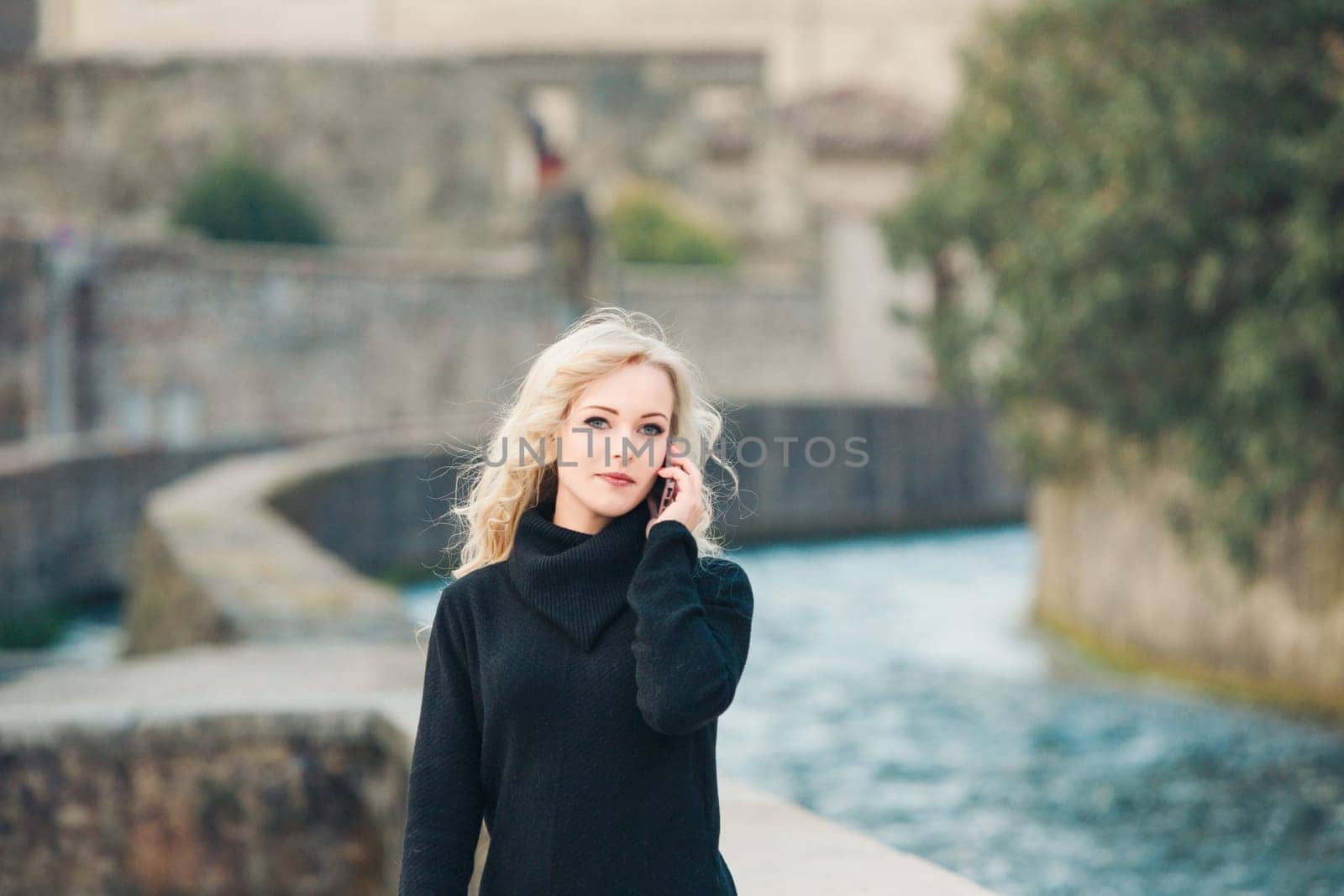 Young woman is enjoying a sunny day in a historic european city, talking on her cellphone as she walks beside a river