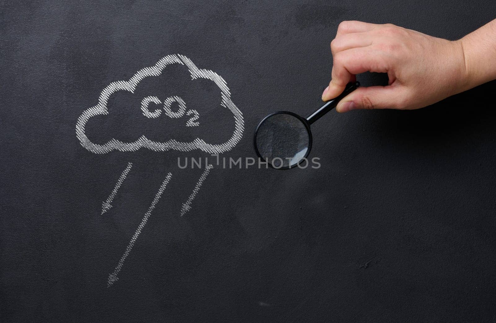Female hand holds a magnifying glass against a black chalkboard background and a cloud, concept of reducing CO2 emissions