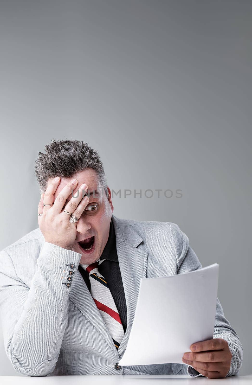 Businessman is sitting at a desk holding a document and looking shocked, with his hand over his face and mouth wide open