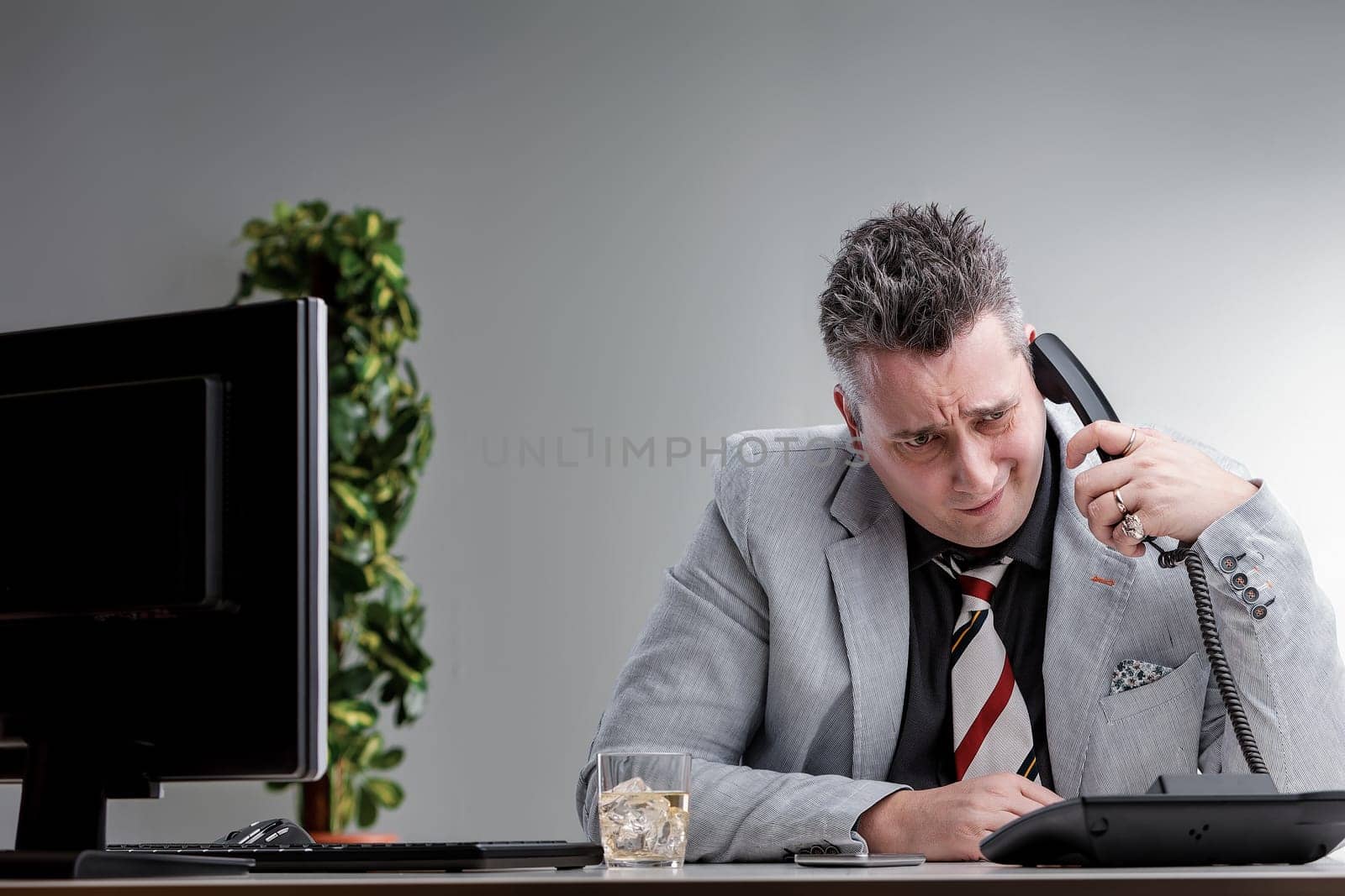 Stressed businessman drinking alcohol and pretending to be working while talking on the phone issues of alcoholism in the workplace