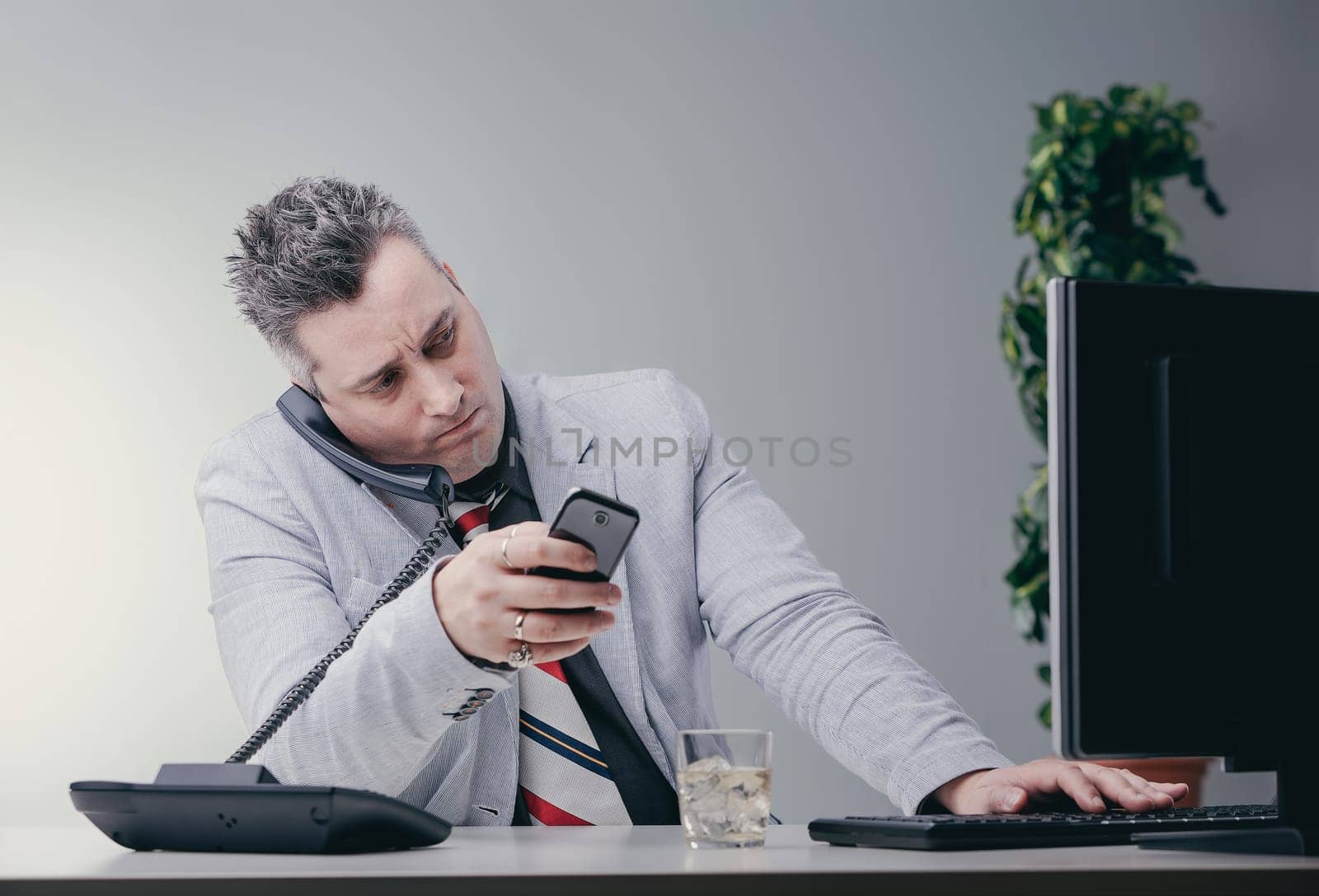 Businessman is struggling to keep up with his workload, looking stressed while using multiple devices at once in a modern office