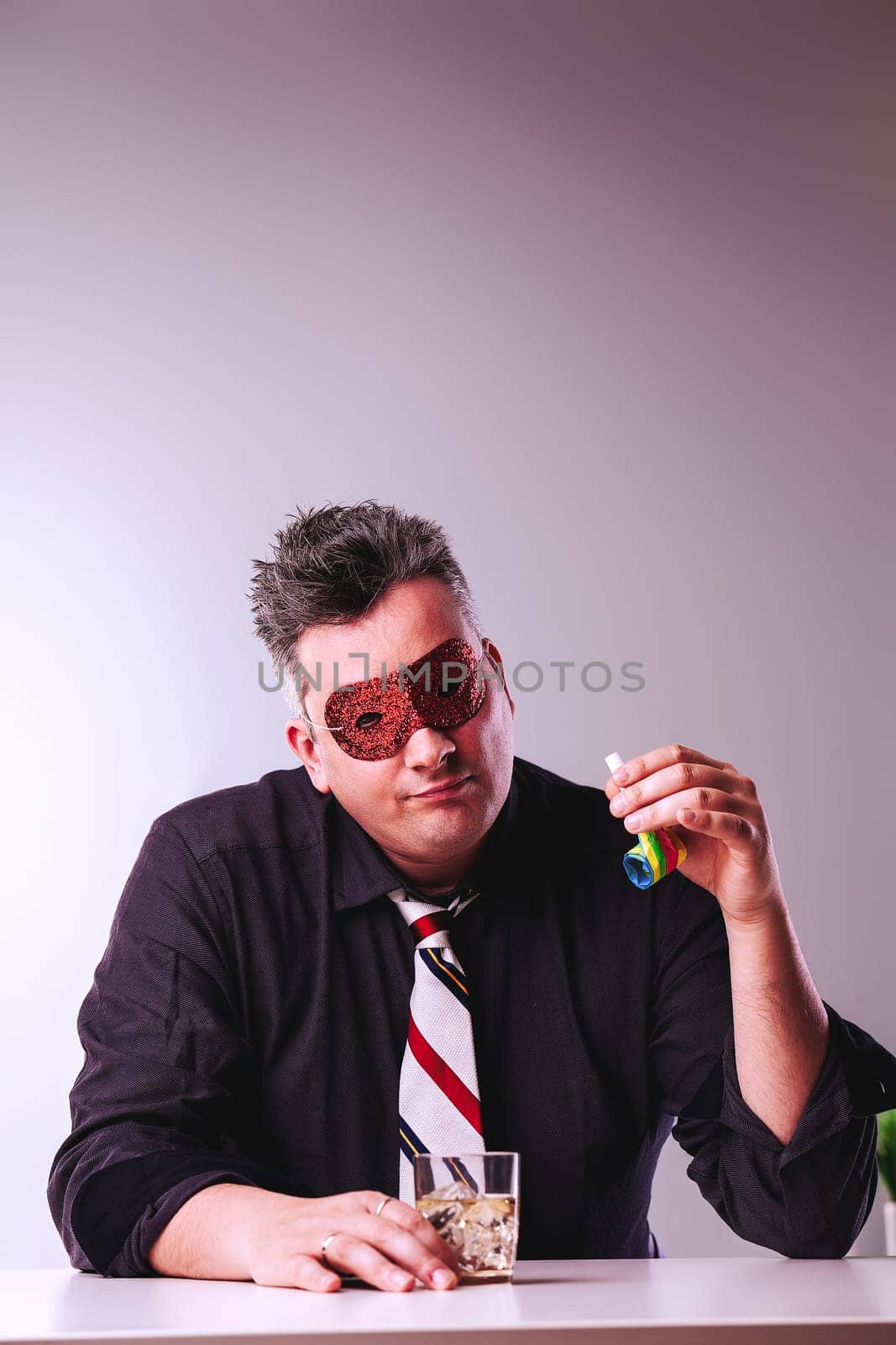 Sad man in a suit sits alone in an office, wearing a party mask and sipping whiskey, embodying loneliness and burnout