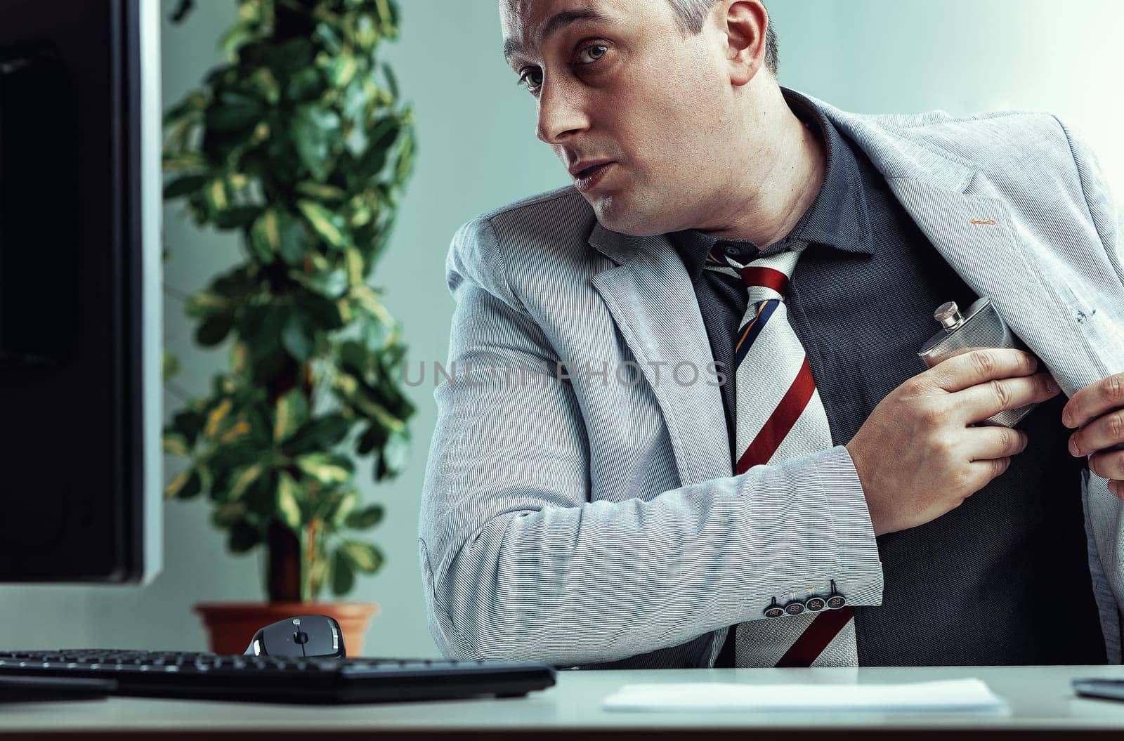 Businessman looking scared while taking a hip flask out of his jacket at work, hiding alcohol addiction issues of alcoholism in the workplace