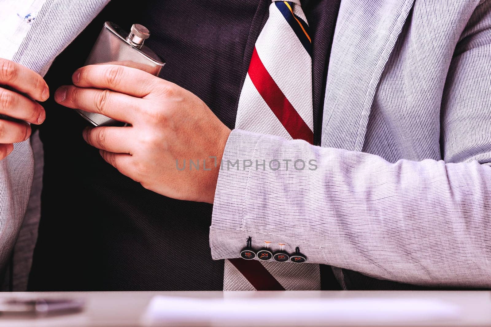 Businessman discreetly hides a flask in his suit pocket, coping with work stress through alcohol. His hand slips the bottle in, showing his unhealthy dependence