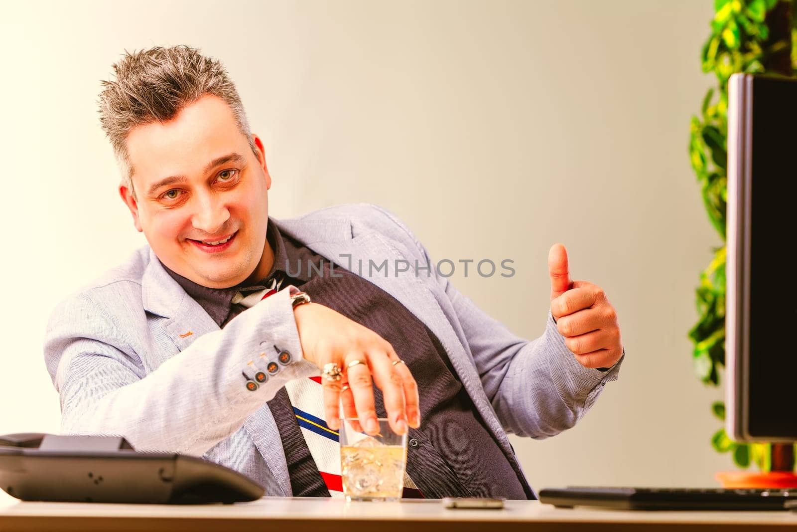 Businessman is sitting at his desk, drinking alcohol and giving a thumbs up. He looks happy and satisfied with his work