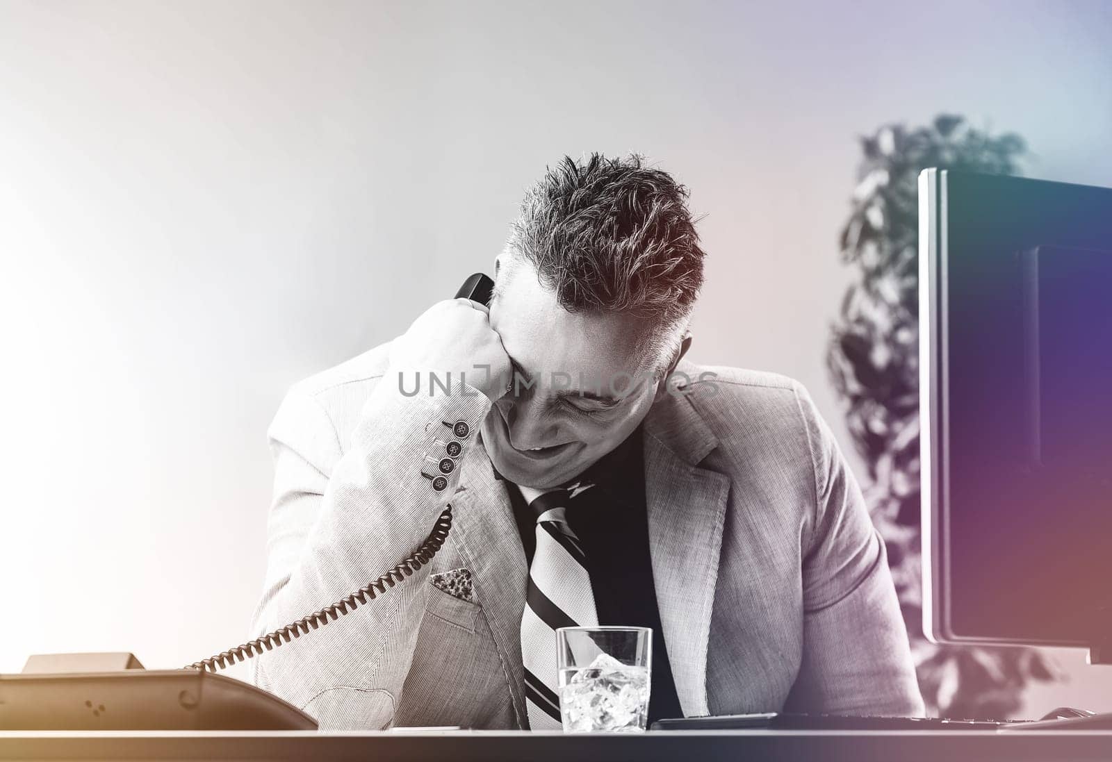 Businessman is laughing during a phone call at his desk while hiding his alcoholism and inner pain issues of alcoholism in the workplace