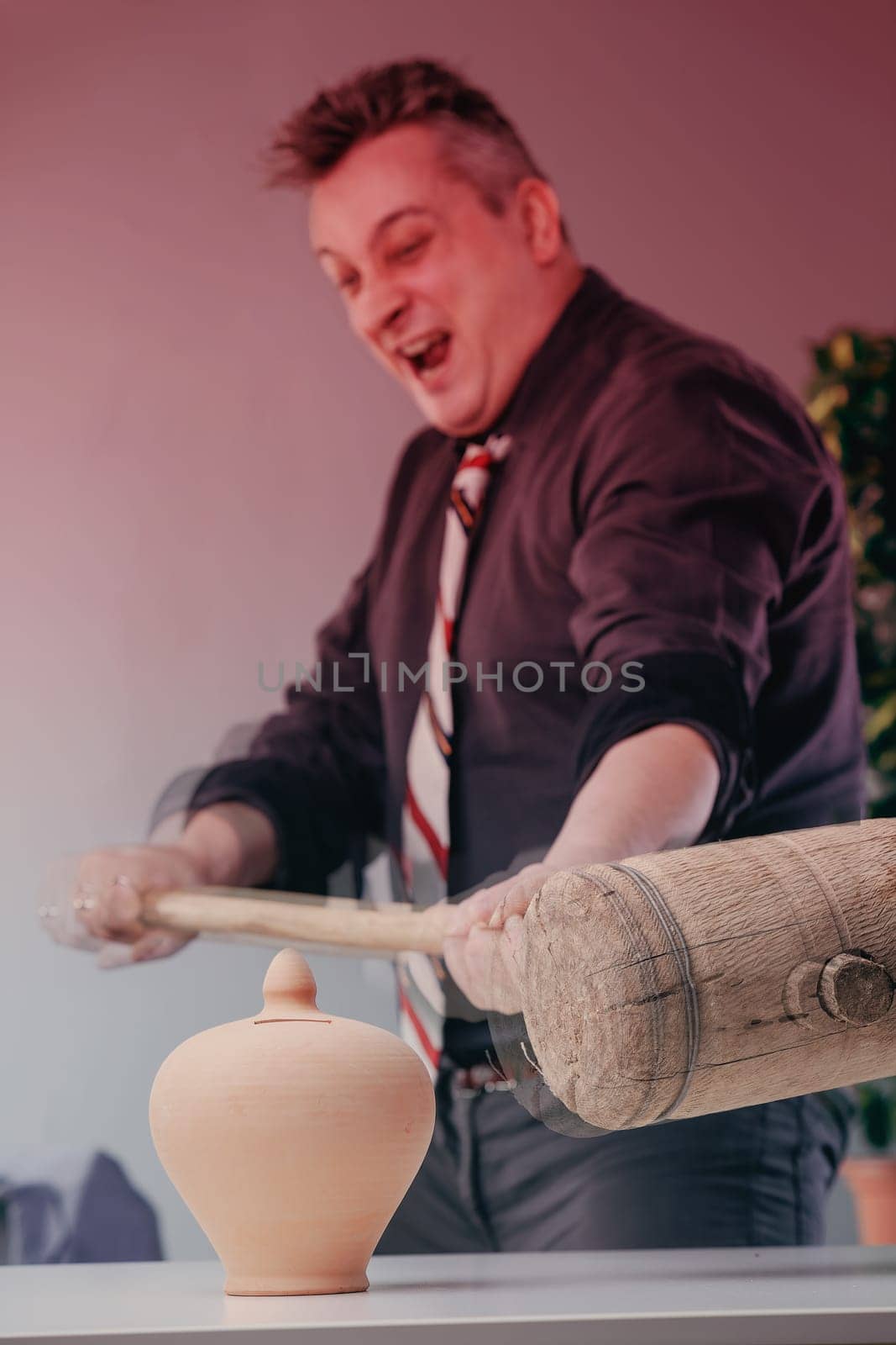 Furious businessman laughing hysterically is about to break a piggy bank with a large mallet, symbolizing his rage and frustration