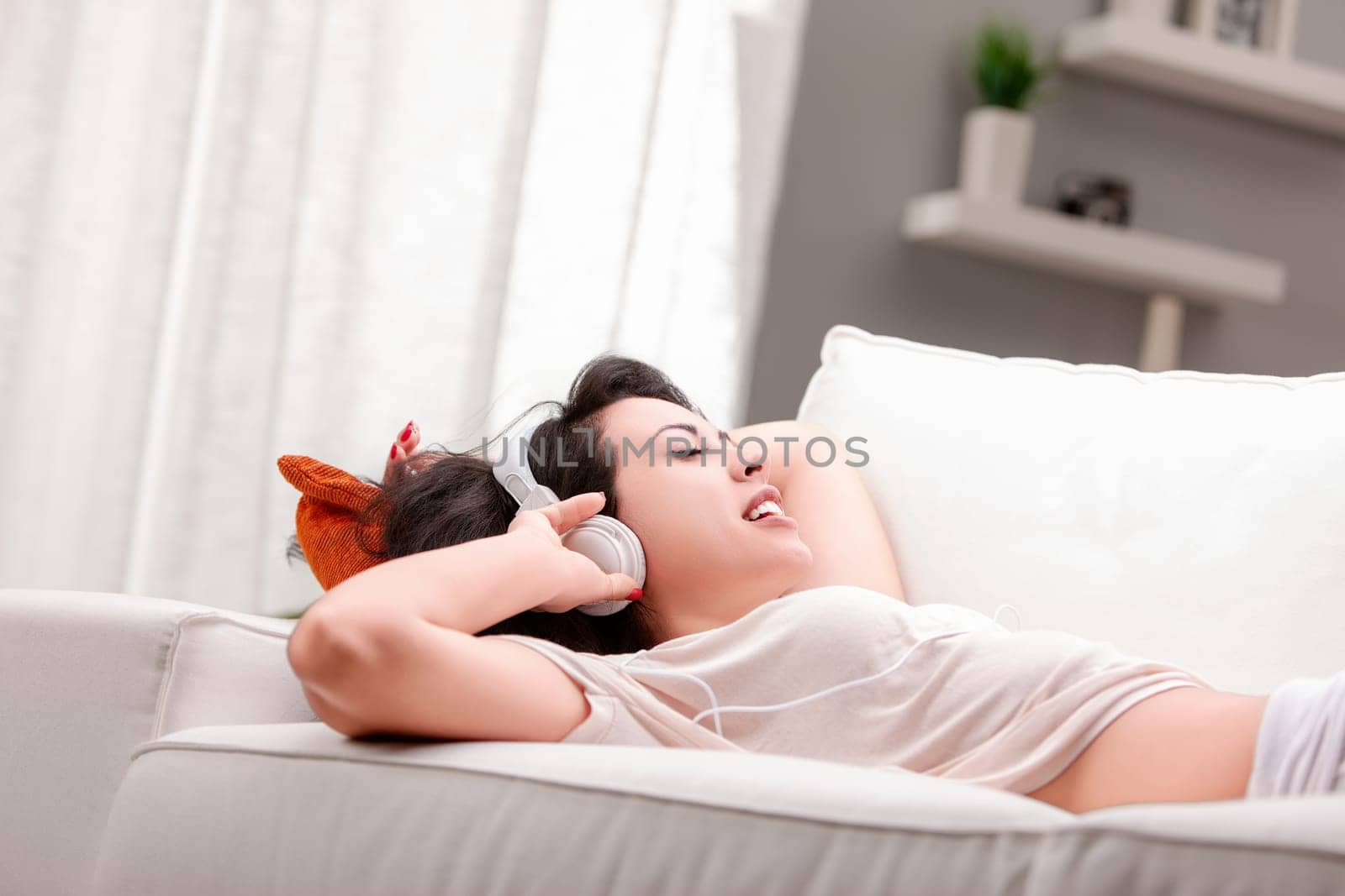 Young woman is lying on a white sofa with her eyes closed, enjoying music through her headphones. She is wearing casual clothes and appears to be very relaxed