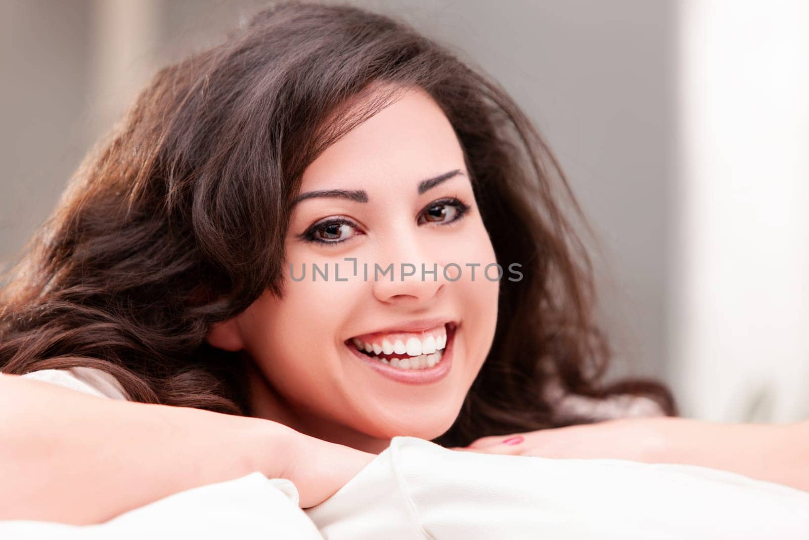 Young woman with long brown hair is smiling while relaxing on a white sofa in her living room, enjoying a peaceful moment at home