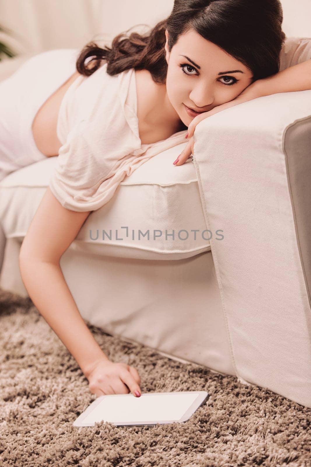 Lifestyle A young woman is lying on a sofa reaching down to use a digital tablet on the floor