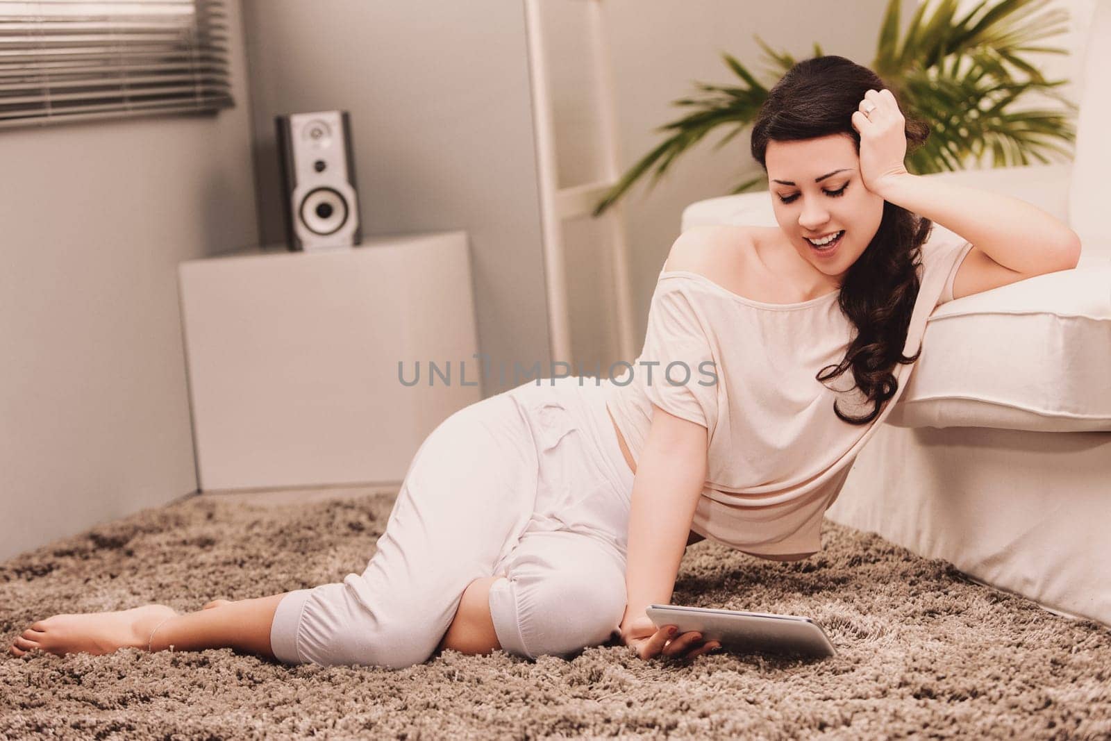 Lifestyle Young woman lying on a rug in her living room using a tablet and smiling, enjoying her free time at home