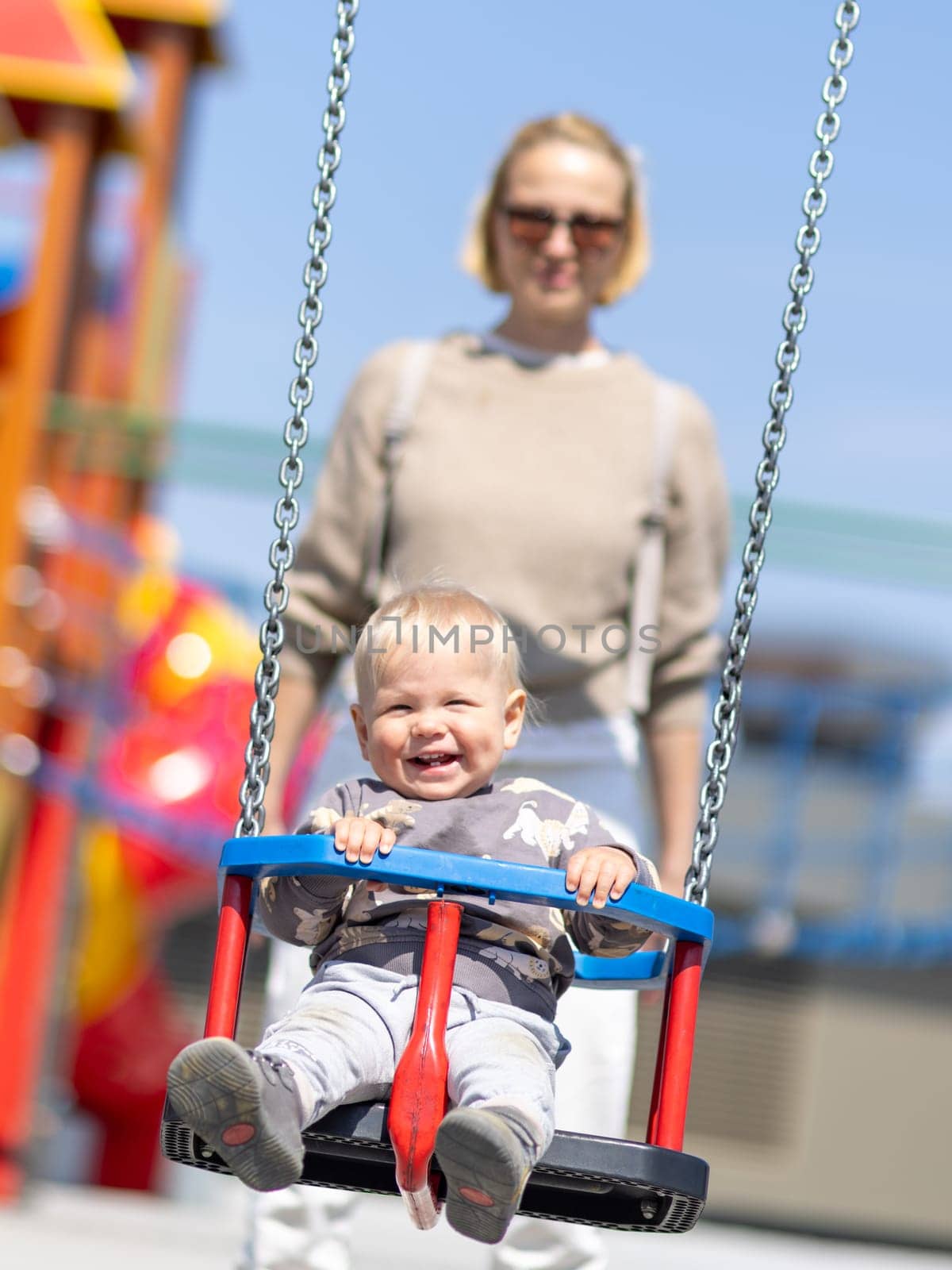 Mother pushing her infant baby boy child on a swing on playground outdoors