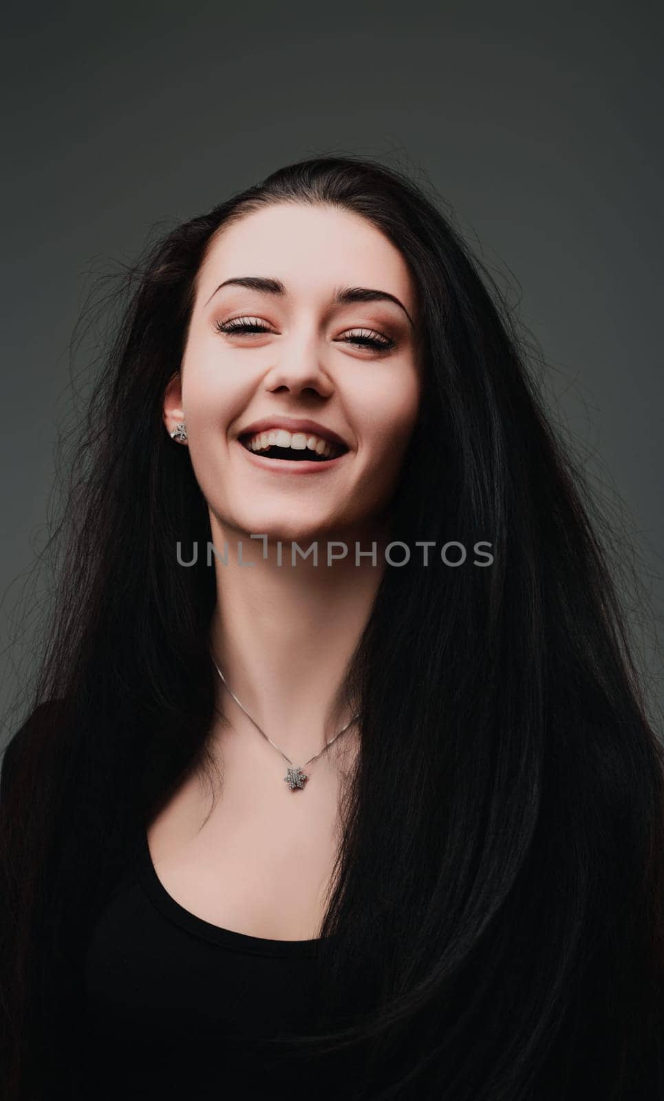Happy brunette woman in a stylish black shirt smiles joyfully in a studio portrait against a grey background, exuding beauty and a carefree vibe