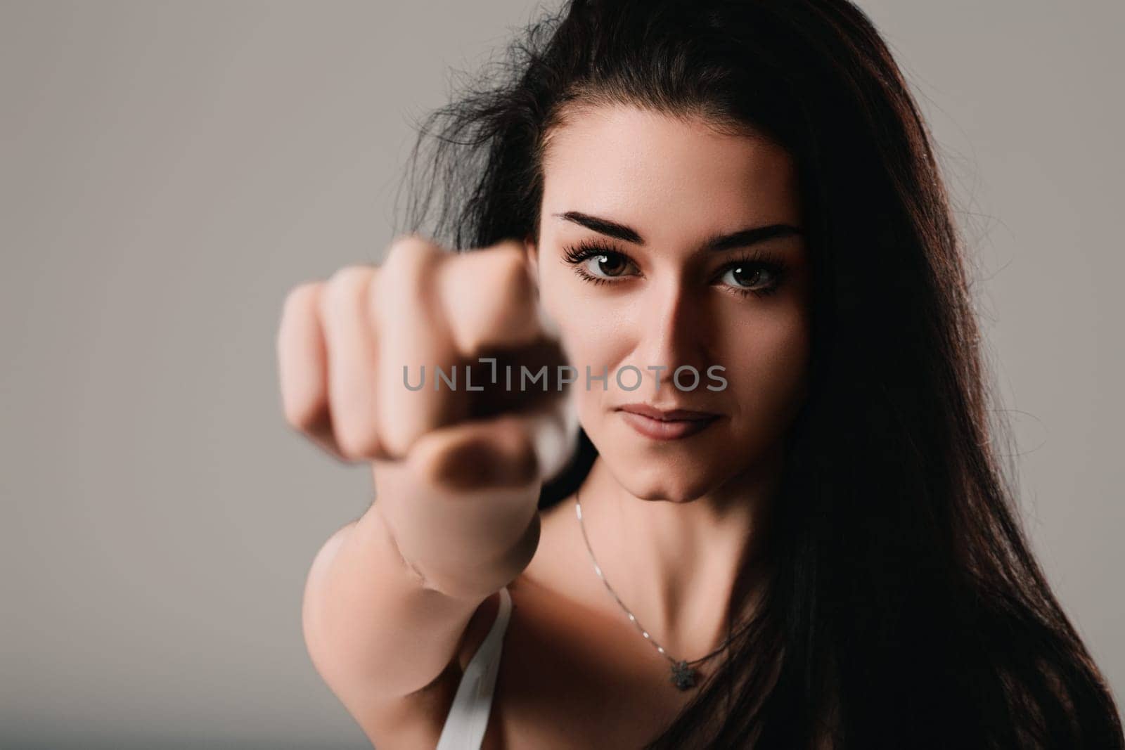 Young woman with dark hair points directly at the camera, looking serious and powerful in a studio shot with a grey background