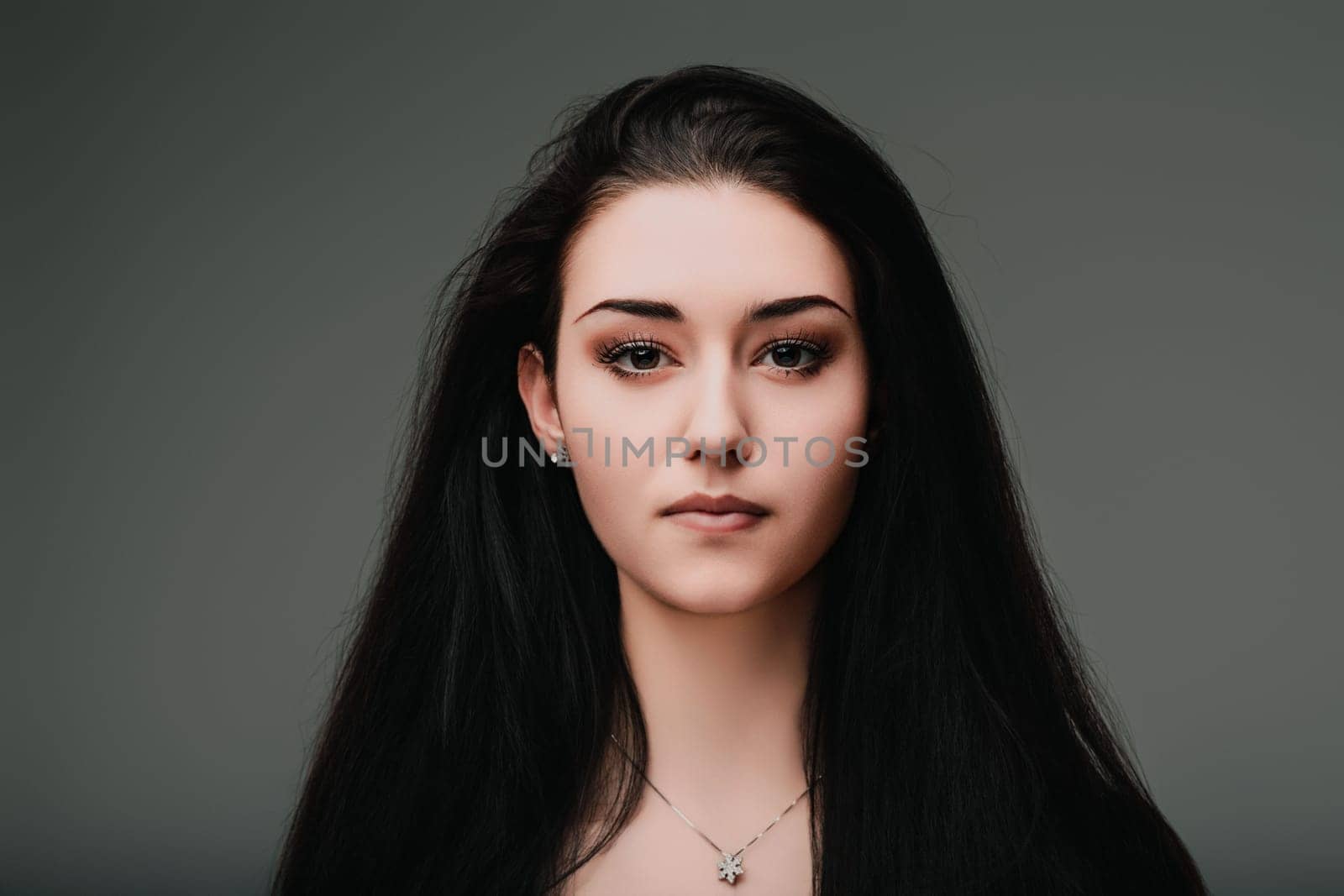 Young woman with long black hair is posing on a gray background, her serious facial expression suggesting deep thought and introspection