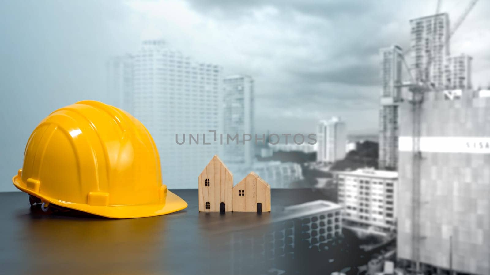Hard hat and wooden house mockup placed at black tabletop on double exposure background of high building and construction site. Project planning presentation, project presentation and project design.