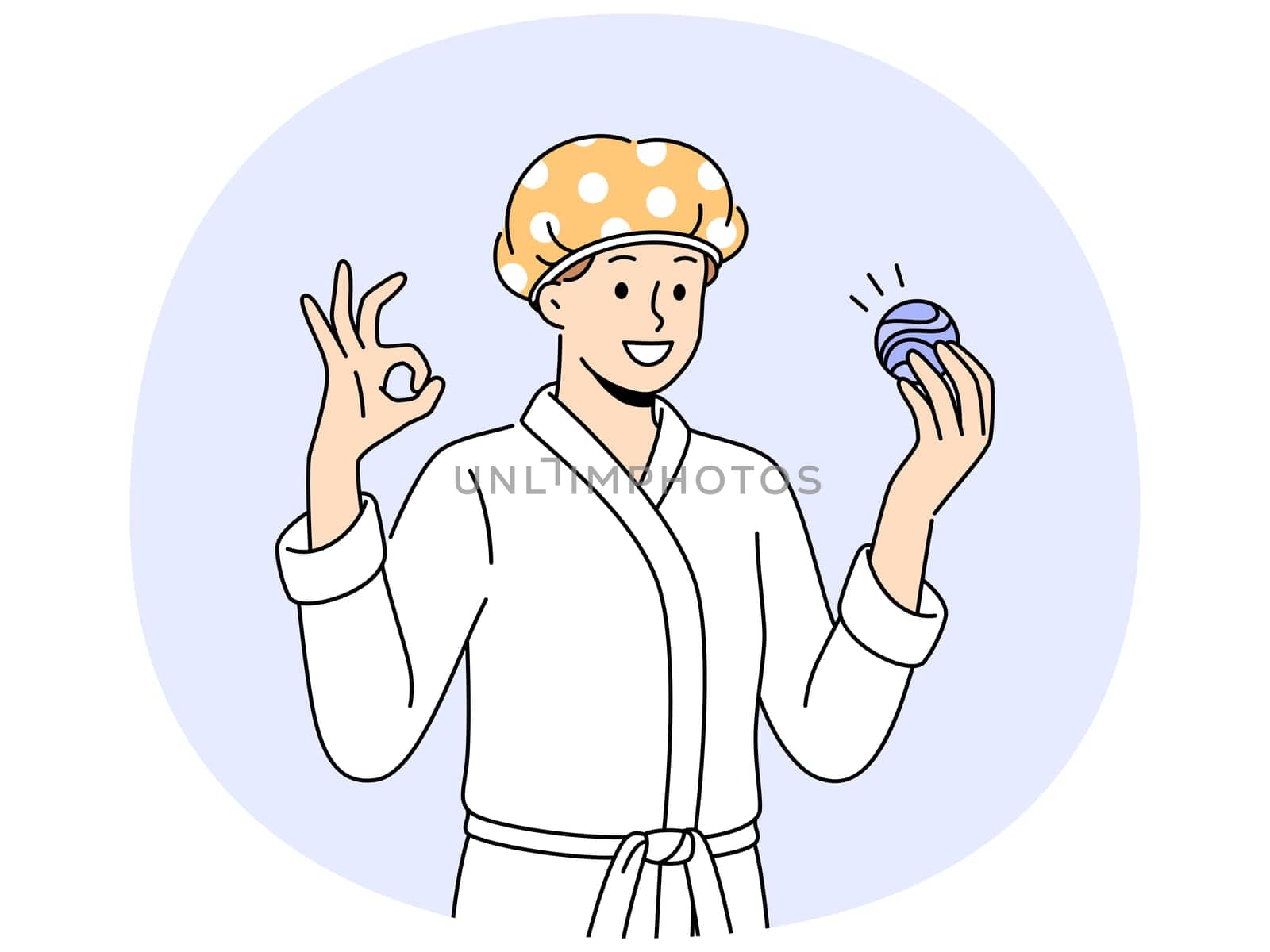 Man with bath bomb is dressed in bathrobe and bandage to protect hair from moisture. Young guy shows OK gesture recommending use of aromatic bath bomb while performing hygiene procedures.