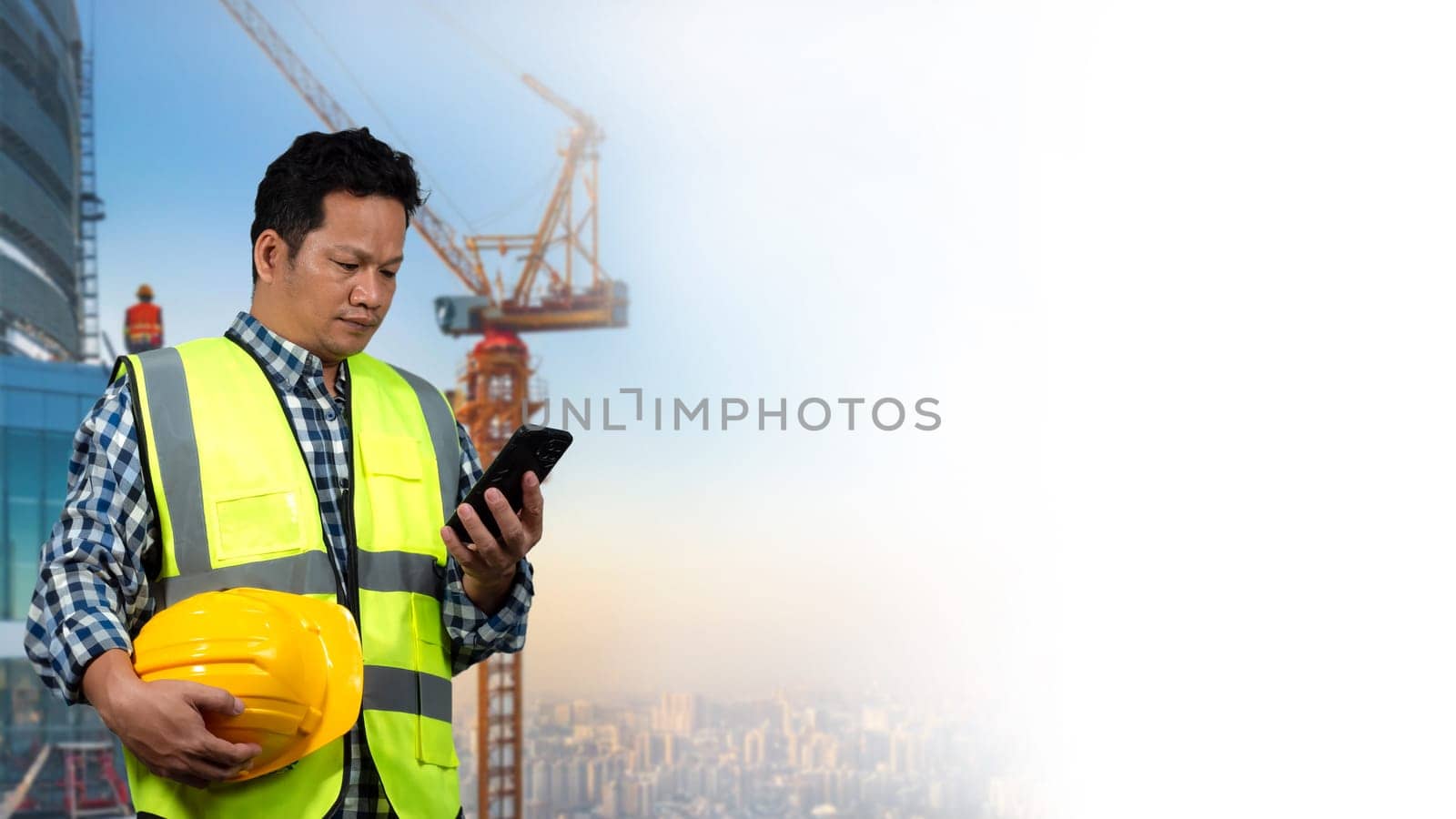 A man wearing a yellow vest and a hard hat is looking at a tablet. The tablet is displaying a construction site with cranes and buildings. The man is focused on the smartphone.