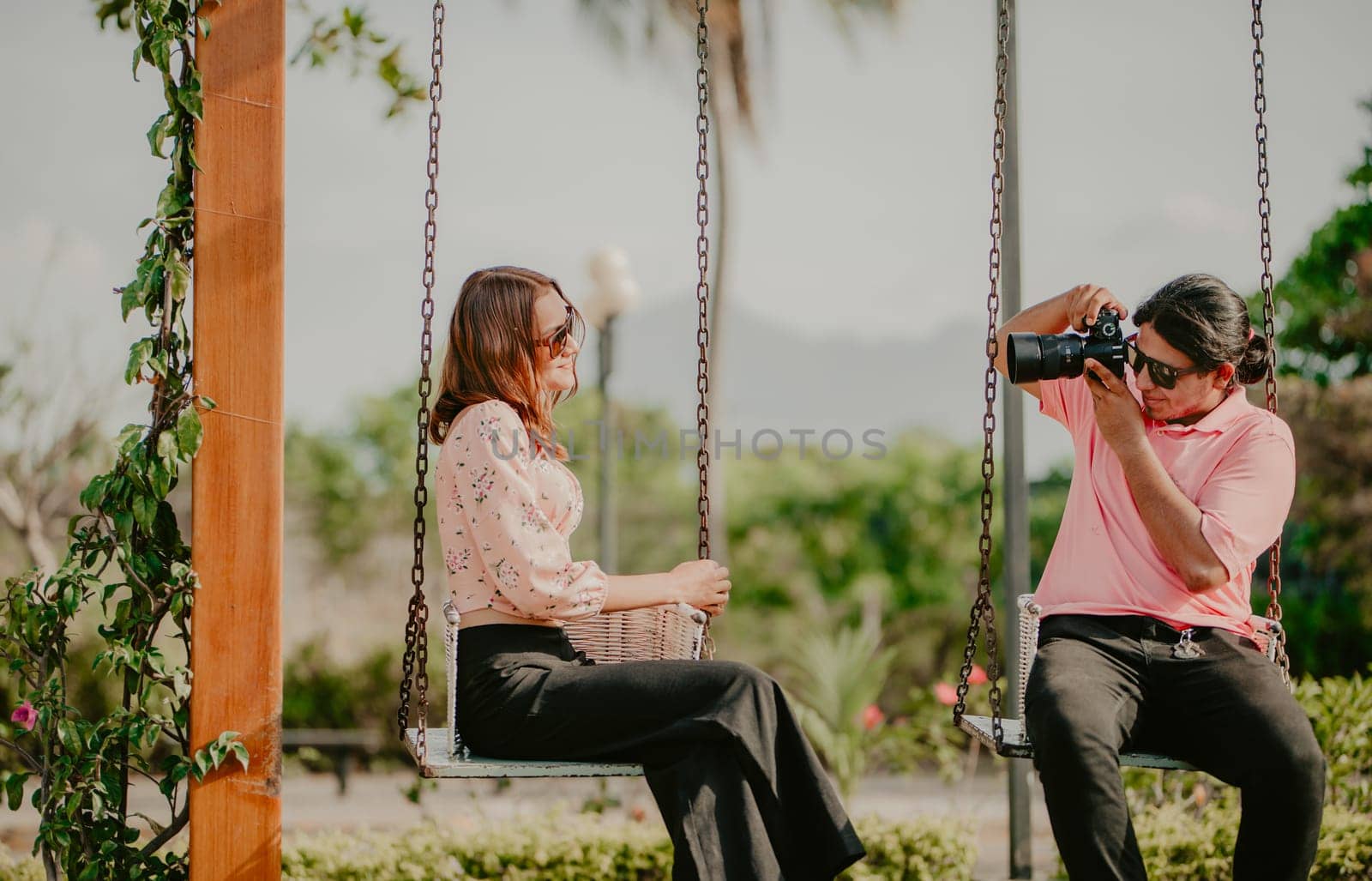 Boyfriend taking a photo of his girlfriend on a swing in a park. Boyfriend photographing girlfriend sitting on a swing