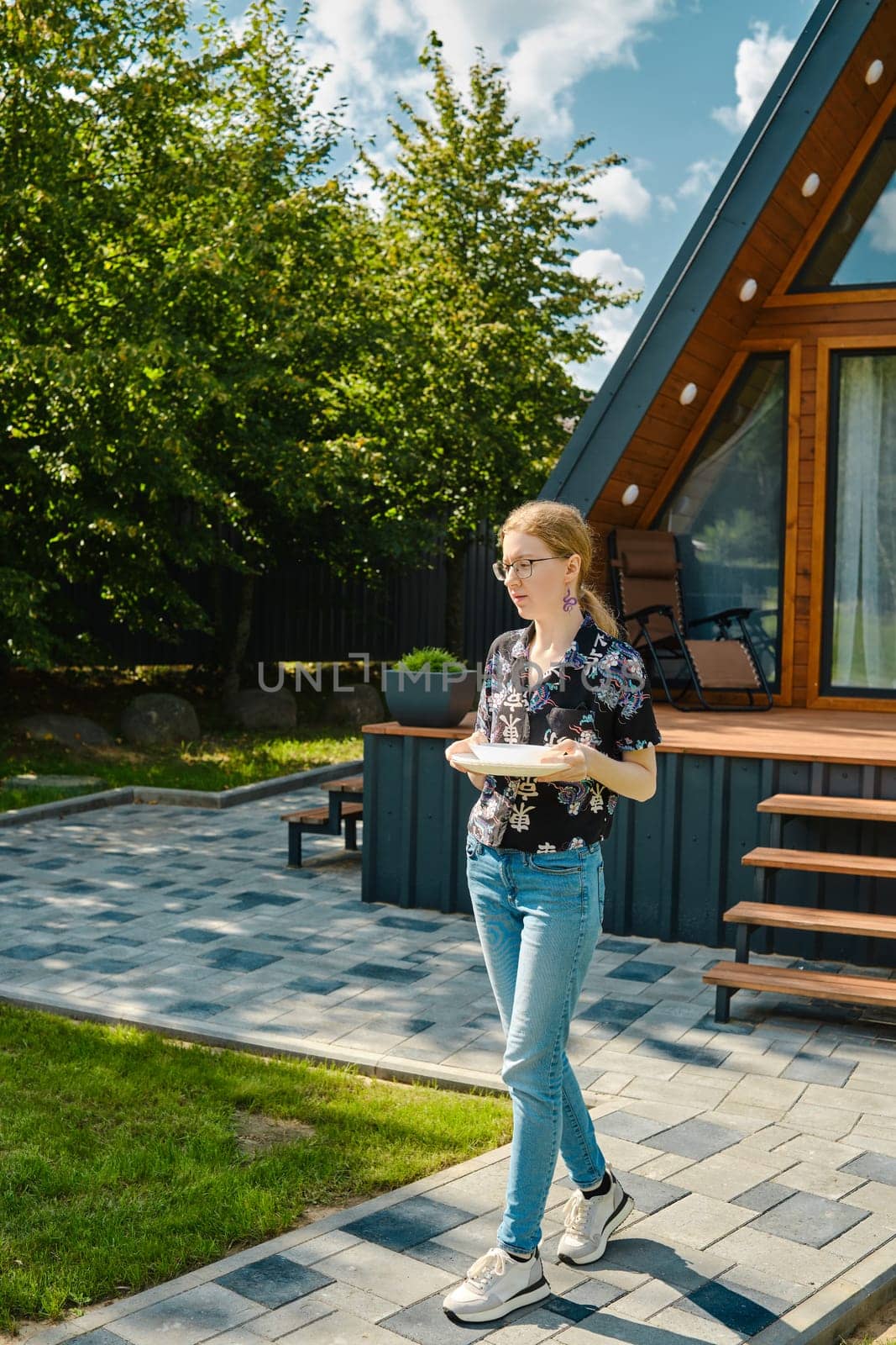 A young woman carries plates to the patio table to lay out grilled food on backyard
