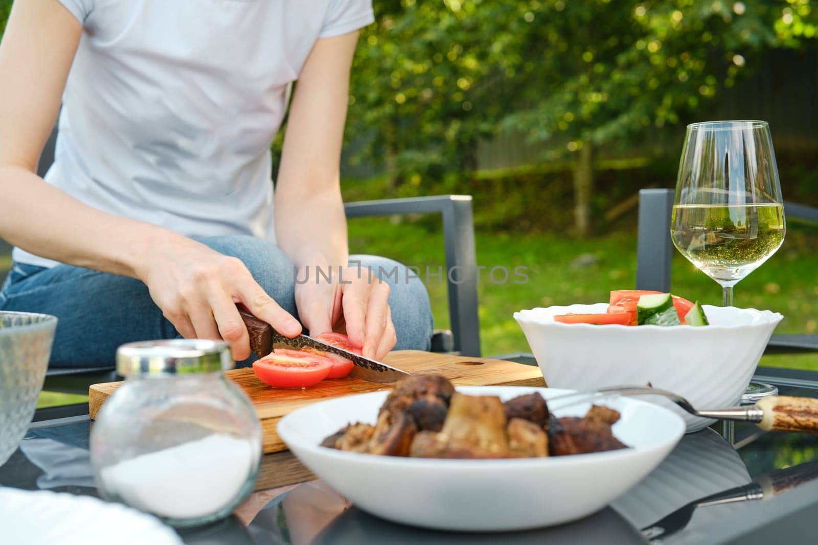 Unrecognizable woman slicing tomato for salad outdoors while sitting in backyard