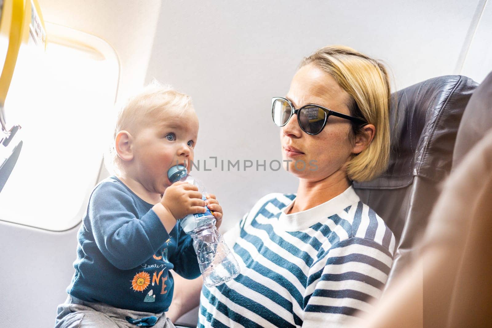 Mom and child flying by plane. Mother holding and playing with her infant baby boy child in her lap during economy comercial flight. Concept photo of air travel with baby. Real people