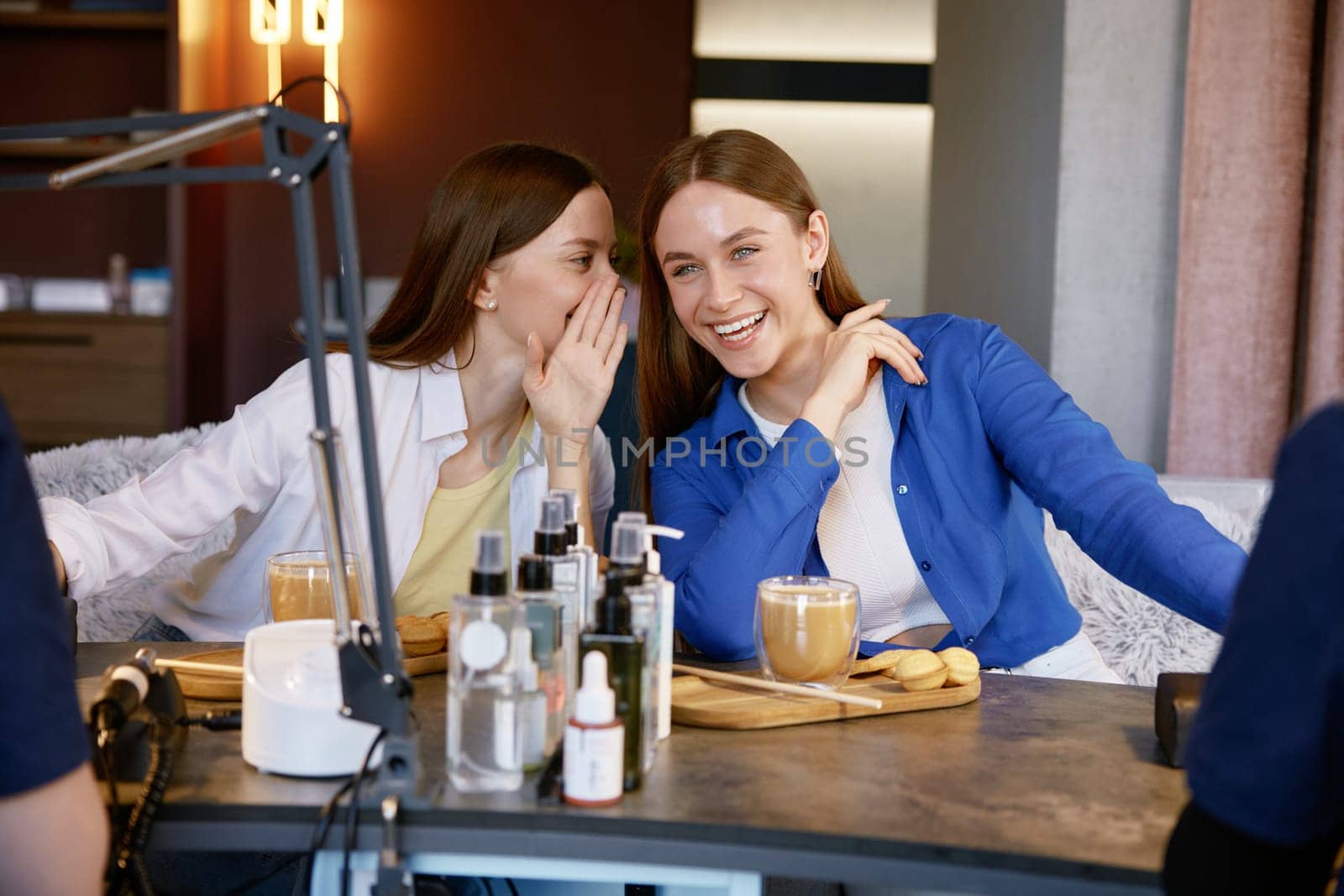 Young woman friends gossiping whispering secrets at nail studio. Two happy girls getting manicure spending fun time together at beauty salon