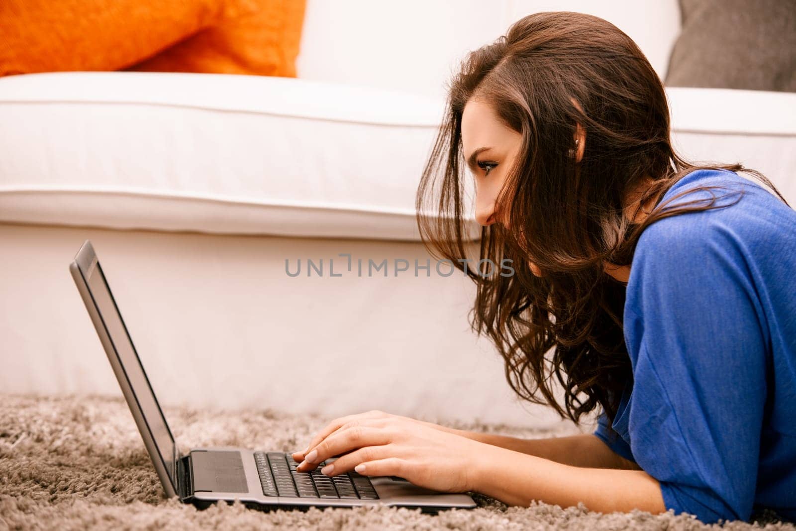 Young woman is lying on her stomach on the carpet and typing on her laptop, she is smiling and seems to be having fun
