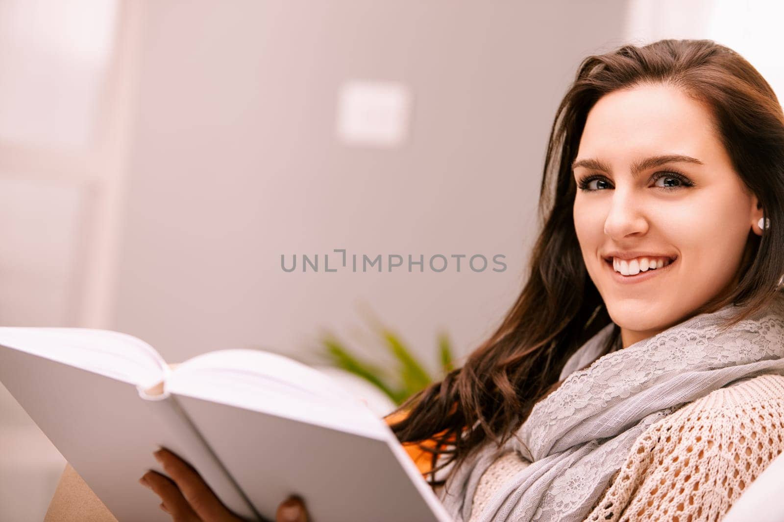 Young woman happily reading a book on a cozy sofa at home, surrounded by warmth and natural light, exuding relaxation and contentment