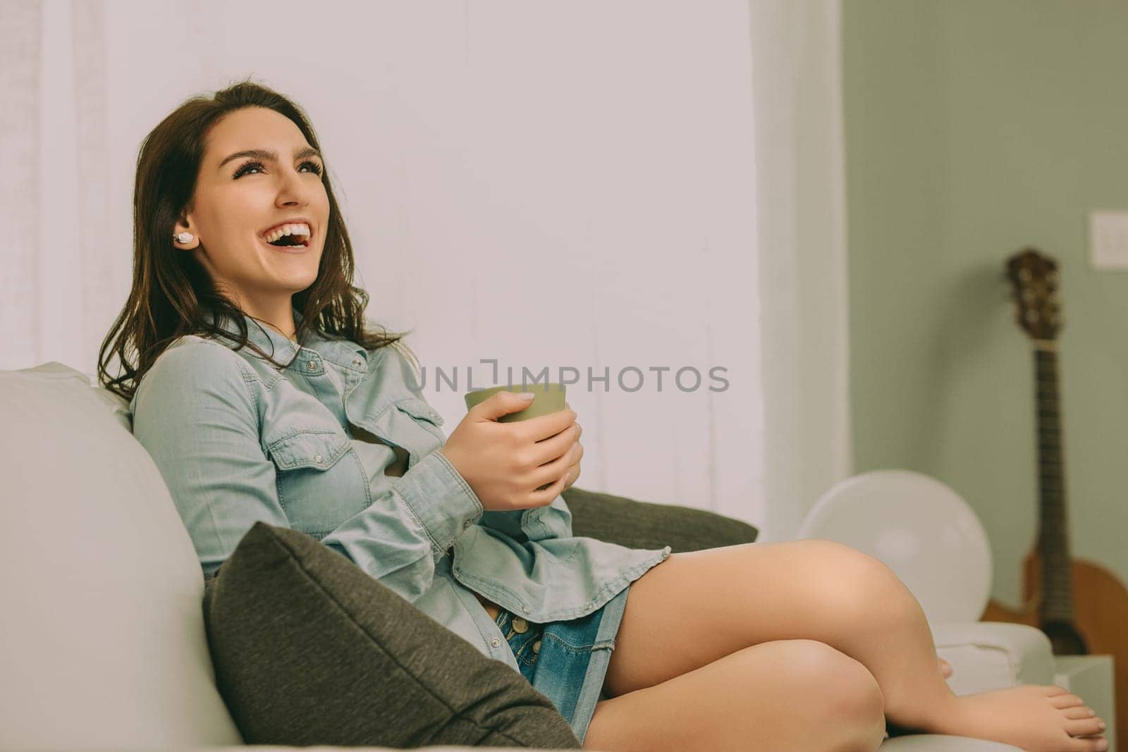 woman with long brown hair sits on a sofa, laughing joyfully as she holds a green mug. Dressed in a light denim shirt and shorts, her relaxed posture reflects a moment of comfort and happiness