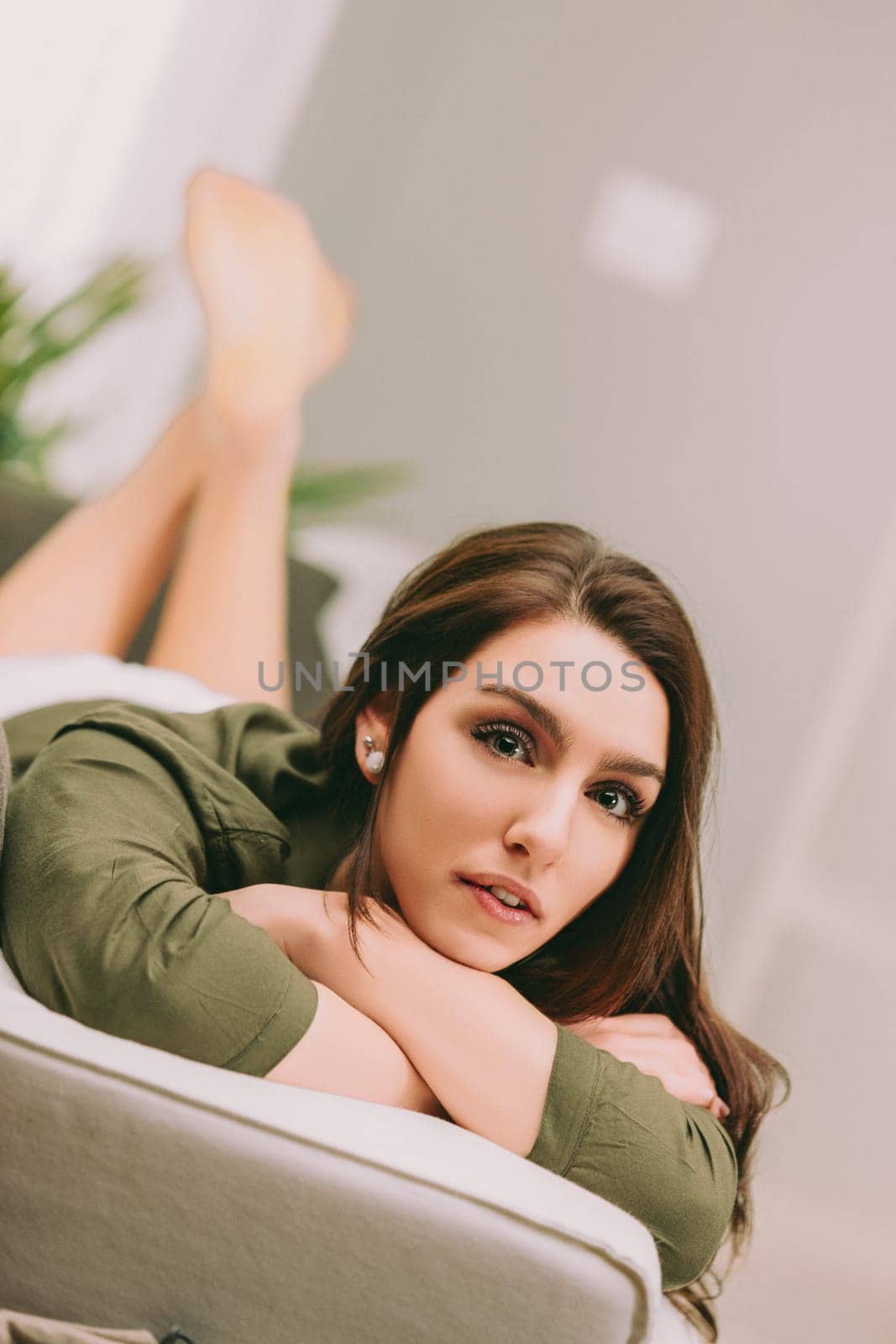 woman, lying on her stomach on a light-colored sofa, rests her head on her arms, looking ahead. Her long brown hair and green shirt blend with the calm, softly lit environment