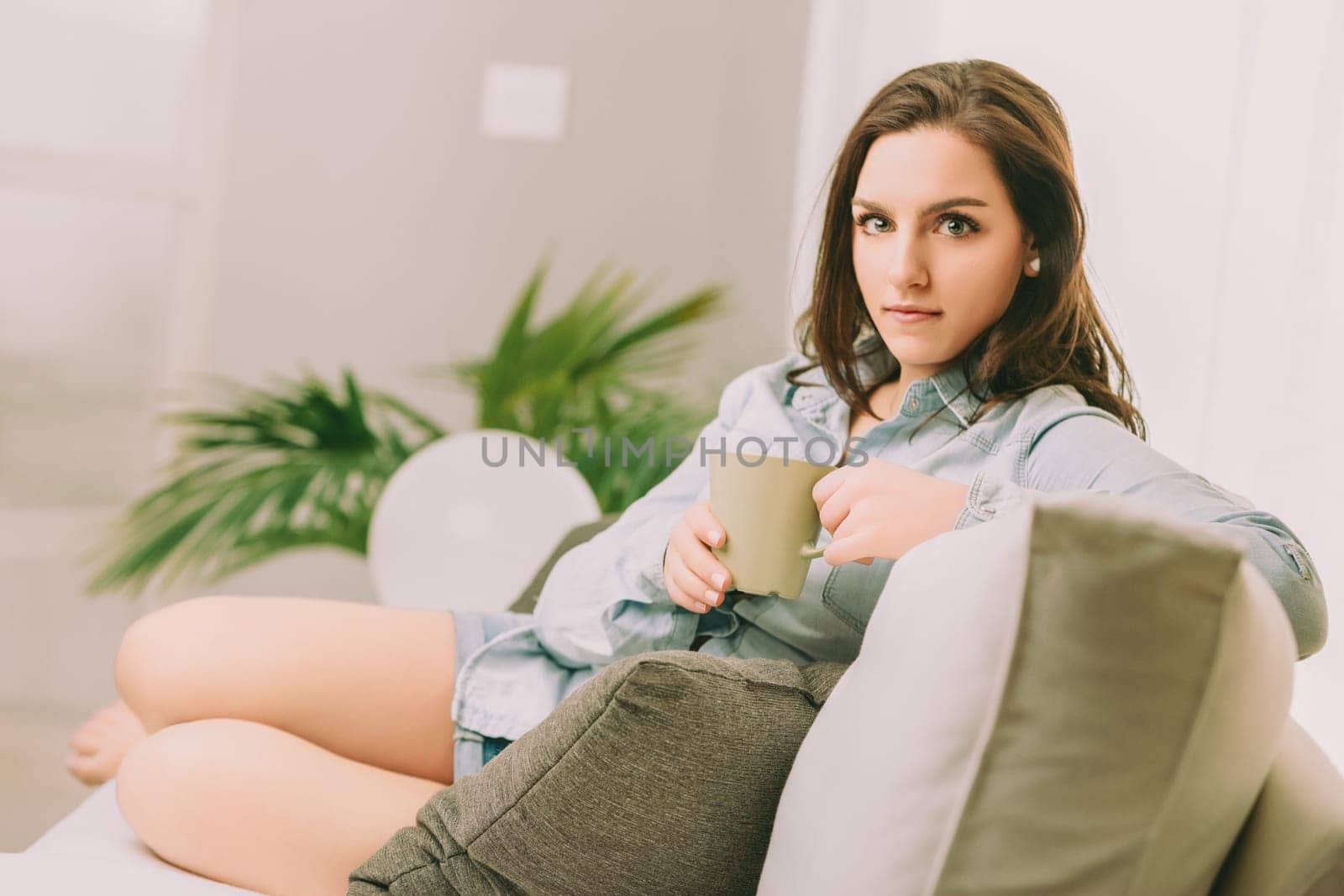 Sitting on a comfortable sofa, a woman with a pensive look holds a green mug. Her relaxed posture, denim shirt, and the surrounding greenery create a tranquil and contemplative atmosphere