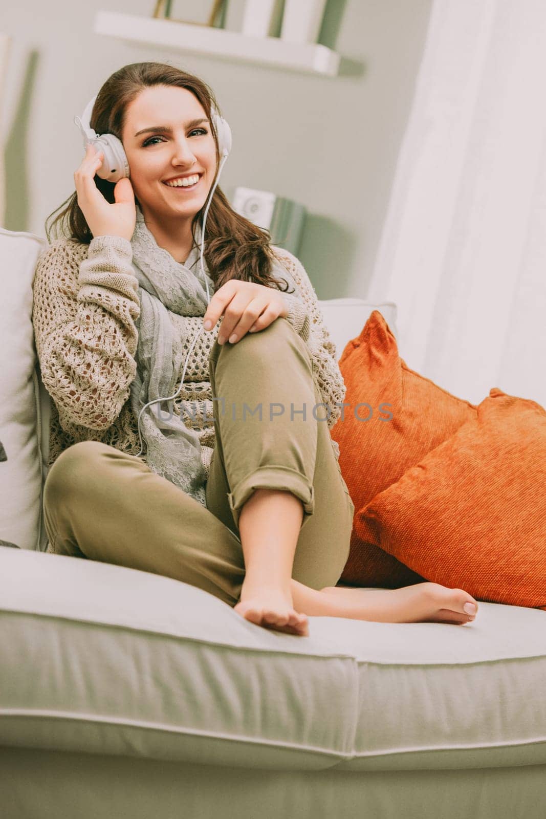 woman with long brown hair sits comfortably on a sofa, smiling as she listens to music through large headphones. Dressed in a cozy sweater and scarf, her relaxed posture adds to the warm, inviting atm