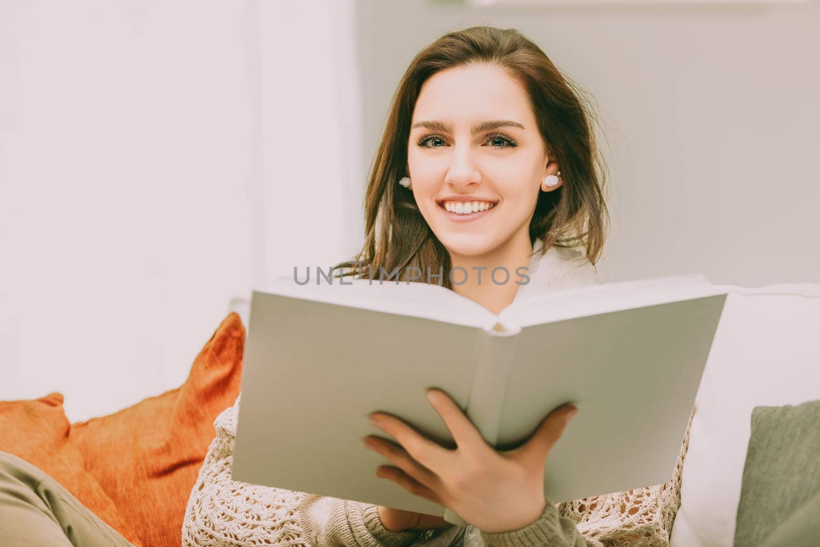 woman with long brown hair sits comfortably, smiling brightly as she reads a book. Dressed in a cozy sweater, her relaxed posture and the bright, inviting setting create a warm and joyful atmosphere