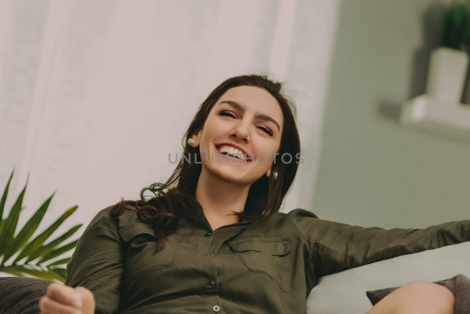 Woman with dark, straight hair smiles broadly while lounging on a modern, light sofa. Dressed casually in a green top and white shorts, her expression radiates warmth and happiness