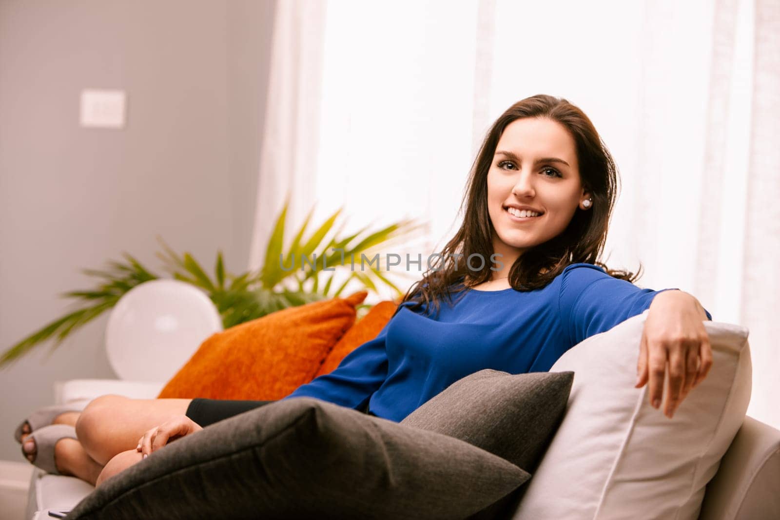 Young woman smiles on her white sofa, enjoying a relaxing day at home in her modern apartment, surrounded by cushions and pillows in a peaceful, happy atmosphere filled with natural light
