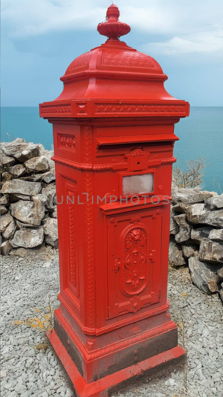 Classic, red British mailbox stands alone on a stone wall, with a view of the wide sea
