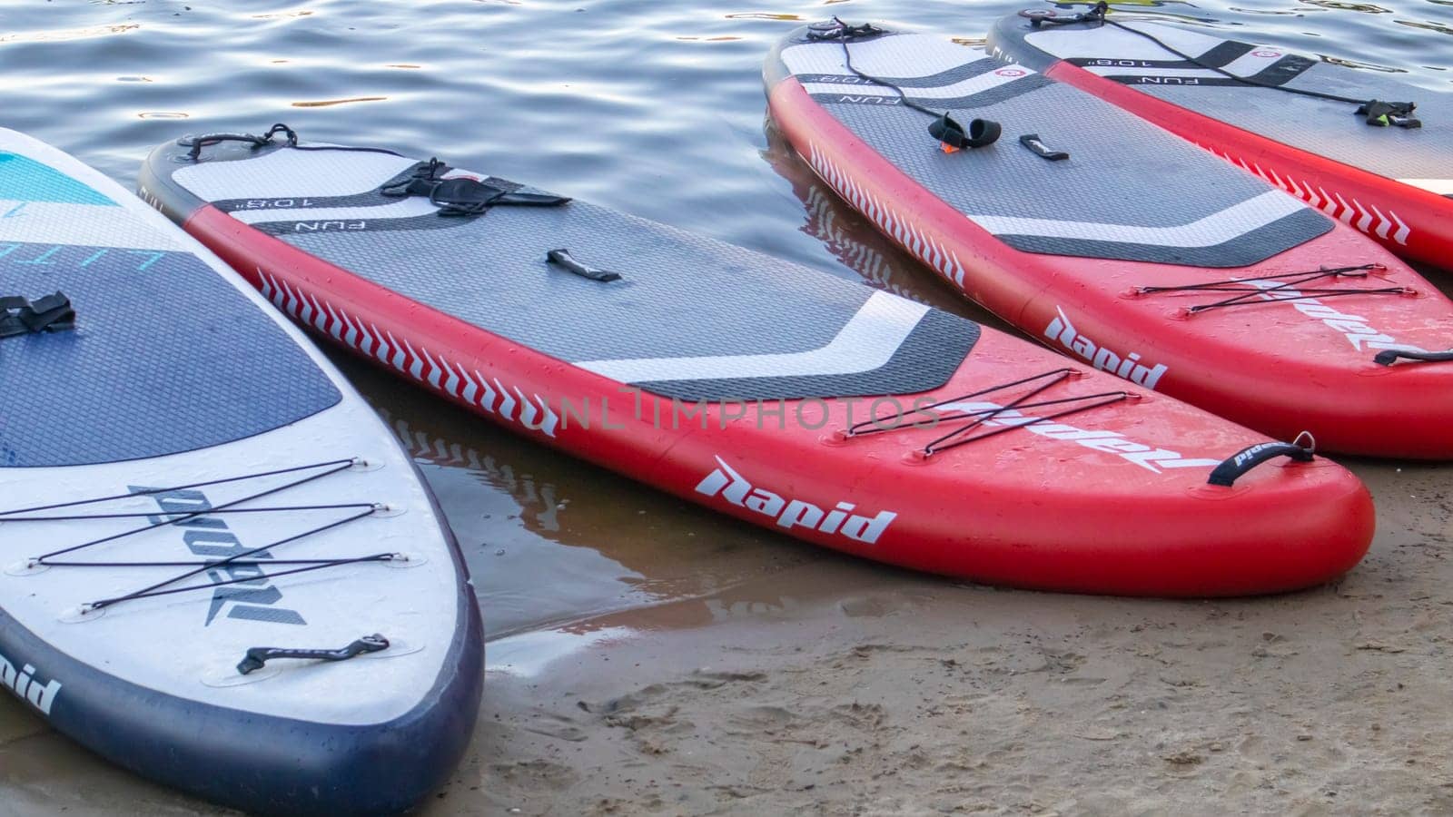 Empty SUP boards sway on the waves on a calm river or lake. Water sports. Lots of boards at the rental shop, the sun reflecting off the ripples of the water. Panning. Ukraine, Kyiv - August 07, 2022