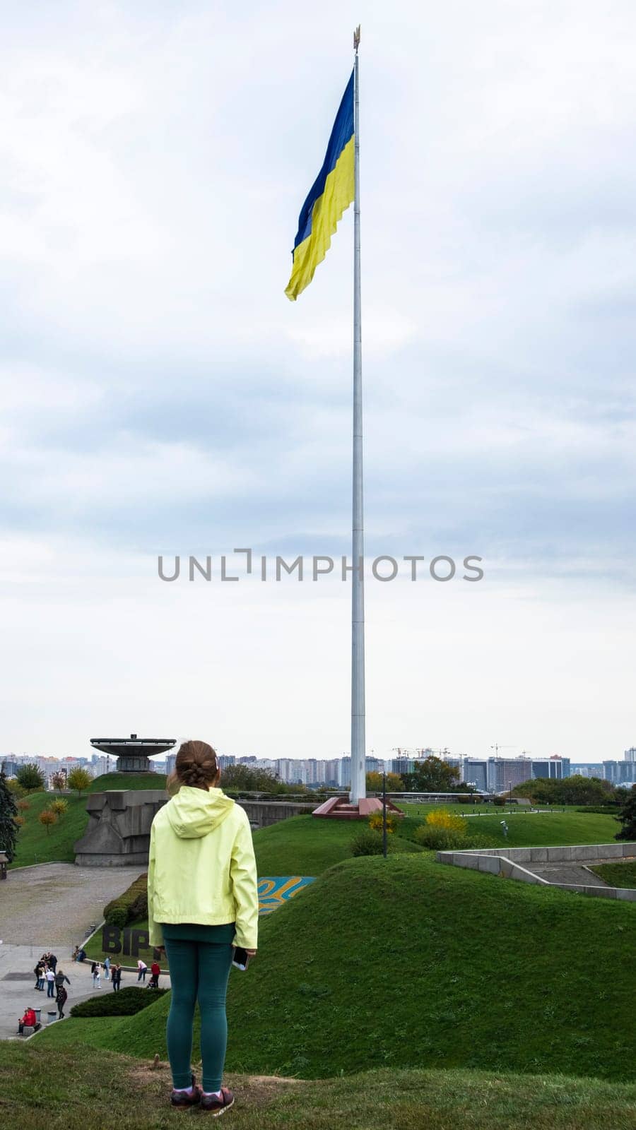 A little girl stands and looks at the flag of Ukraine on a high flagpole. Ukrainians are against the war. Drawing attention to military operations in Ukraine. Ukraine, Kyiv - October 9, 2022