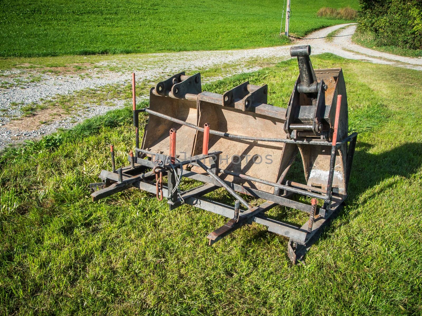 Three excavator buckets of different sizes in a transport frame on a green meadow at the edge of a gravel road fork in Bavaria.