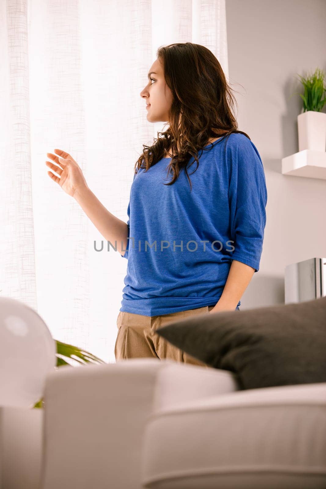 Young woman is standing in her living room, gazing thoughtfully out the window, with a contemplative expression on her face. The soft light creates a serene atmosphere