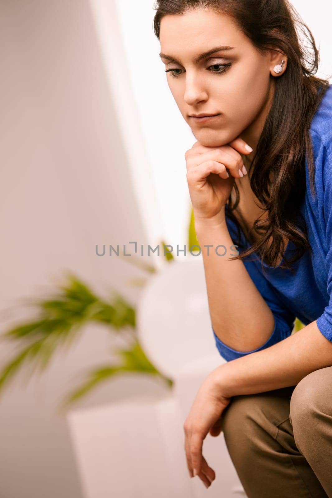 Young woman is sitting on a stool in her living room, resting her chin on her hand and looking down with a worried expression