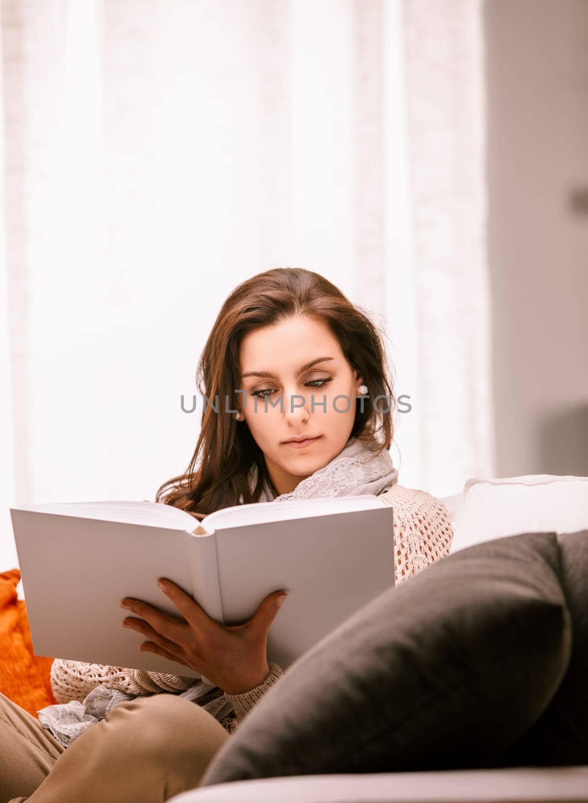 Young woman relaxes on her cozy couch at home, immersed in a captivating book, enjoying a peaceful moment of tranquility