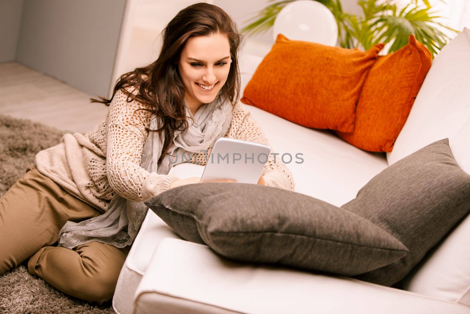Young woman is sitting on the floor in her living room, using a digital tablet and smiling. She is enjoying her free time at home