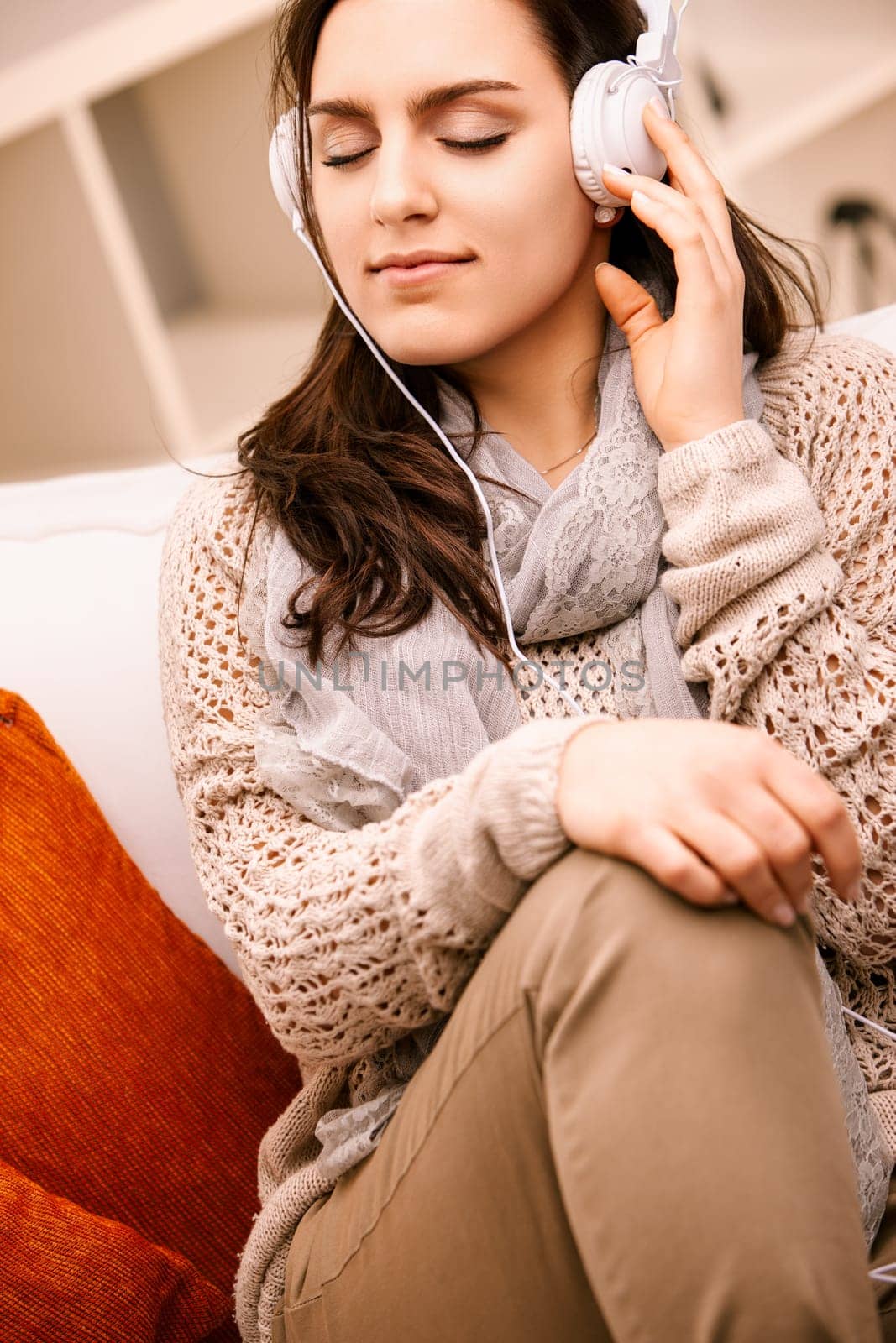 Young woman is enjoying her free time at home listening to music with headphones and relaxing on the couch. She is wearing casual clothing and has her eyes closed