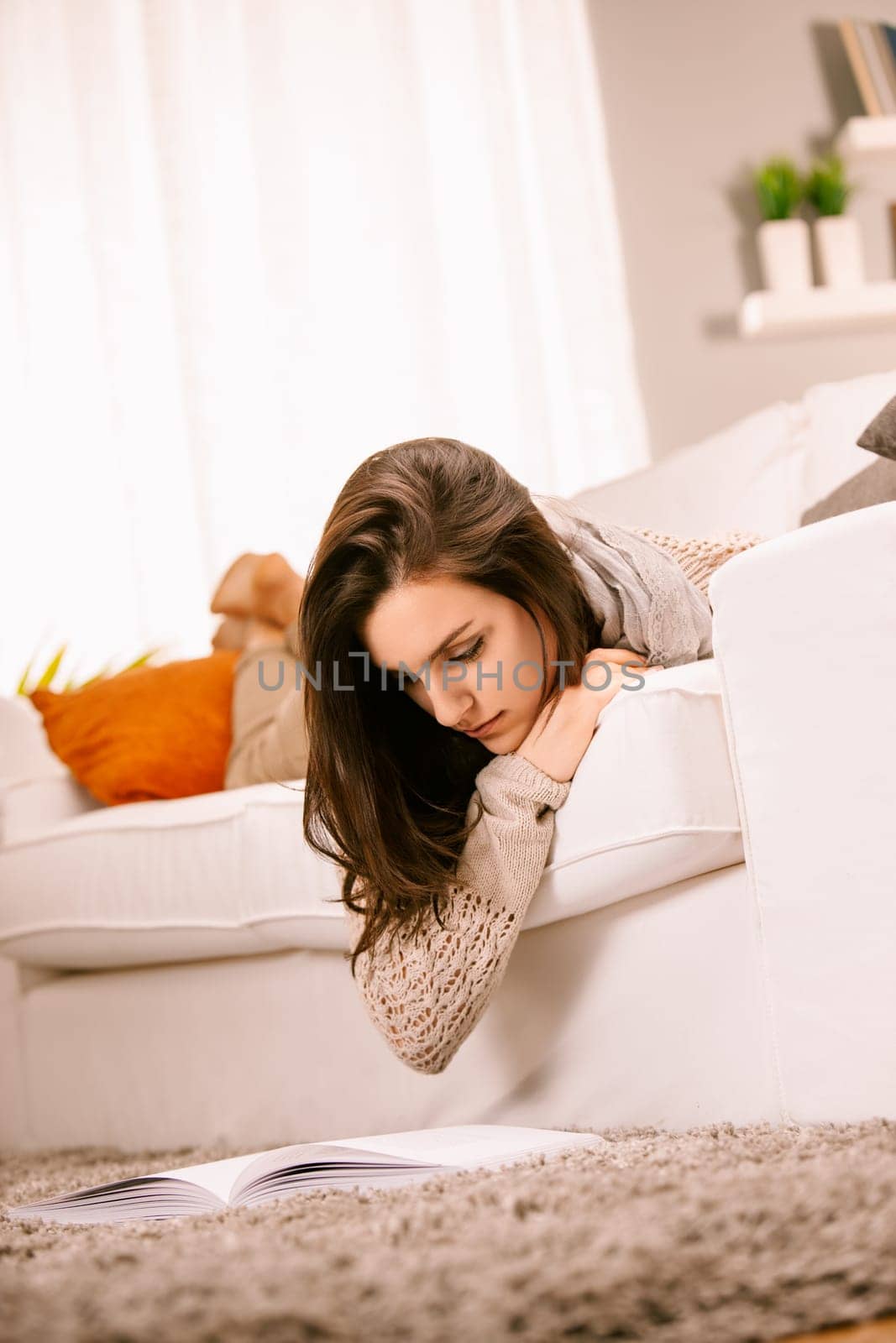 Young woman is lying on her stomach on a white couch with her feet up reading a book on the floor. She is home alone enjoying a relaxing day