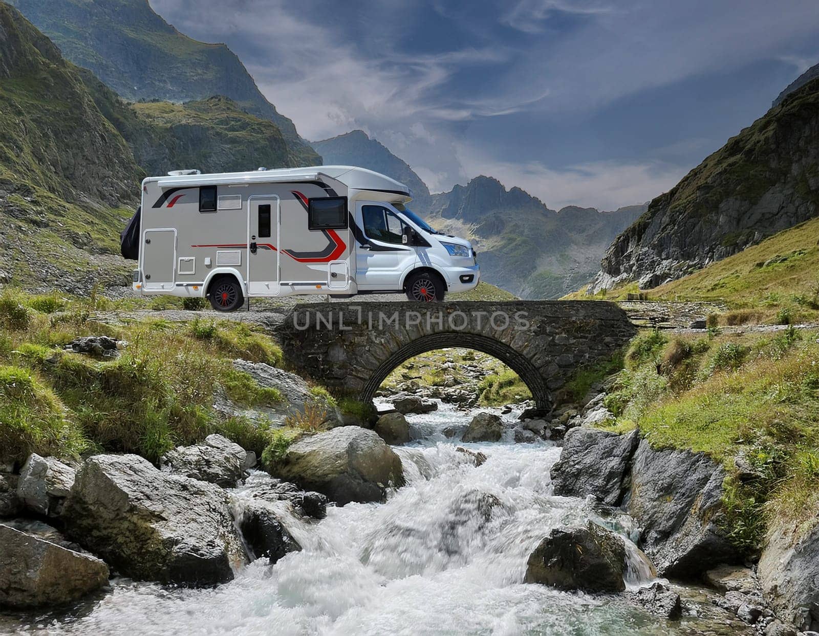 White campervan with red accents, stands on a small bridge over a roaring mountain stream. An impressive mountain backdrop rises in the background.