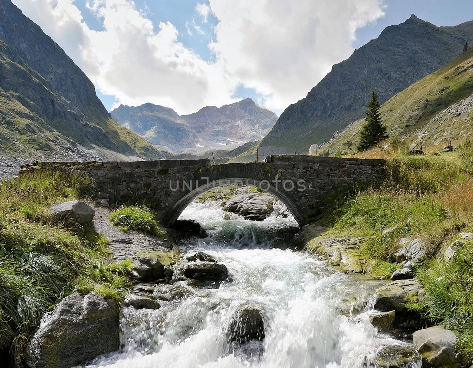 White campervan with red accents, stands on a small bridge over a roaring mountain stream. An impressive mountain backdrop rises in the background.