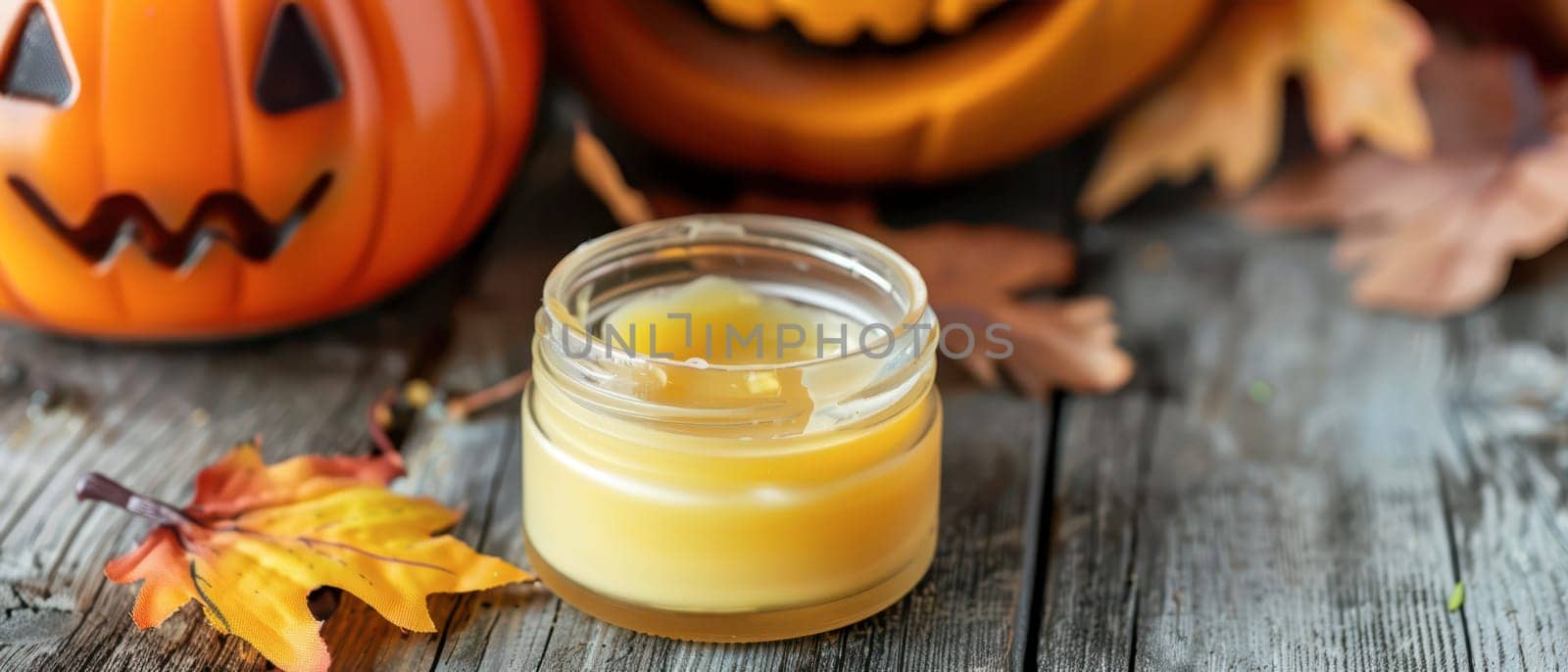 A warm candle sits on a wooden table surrounded by colorful autumn leaves and carved pumpkins, creating a cozy fall atmosphere.