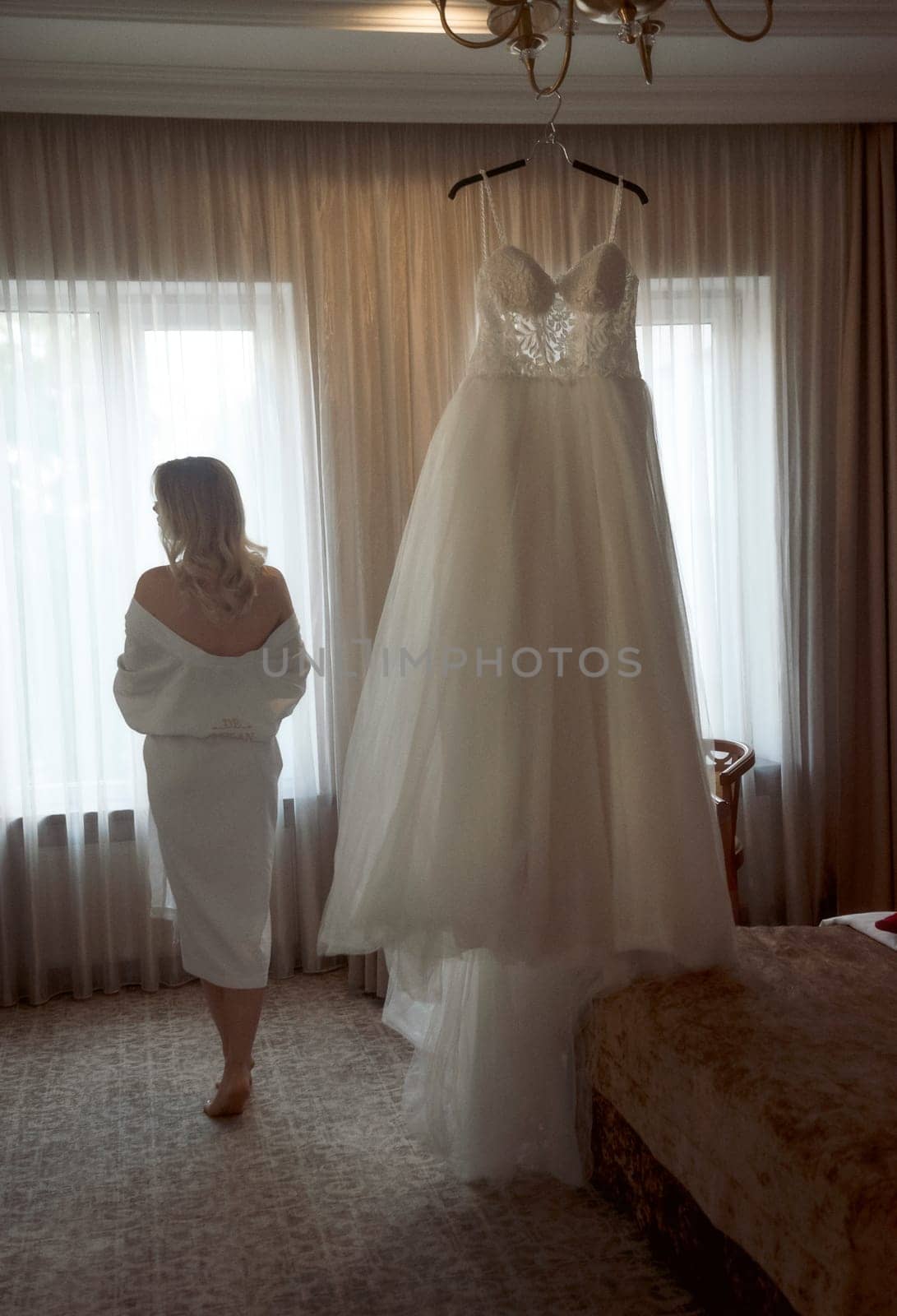 Beautiful girl bride washes her back in the hotel on the wedding morning. Blonde bride stands with her hair done in white robe and gets ready for the wedding day. On the background of a wedding dress